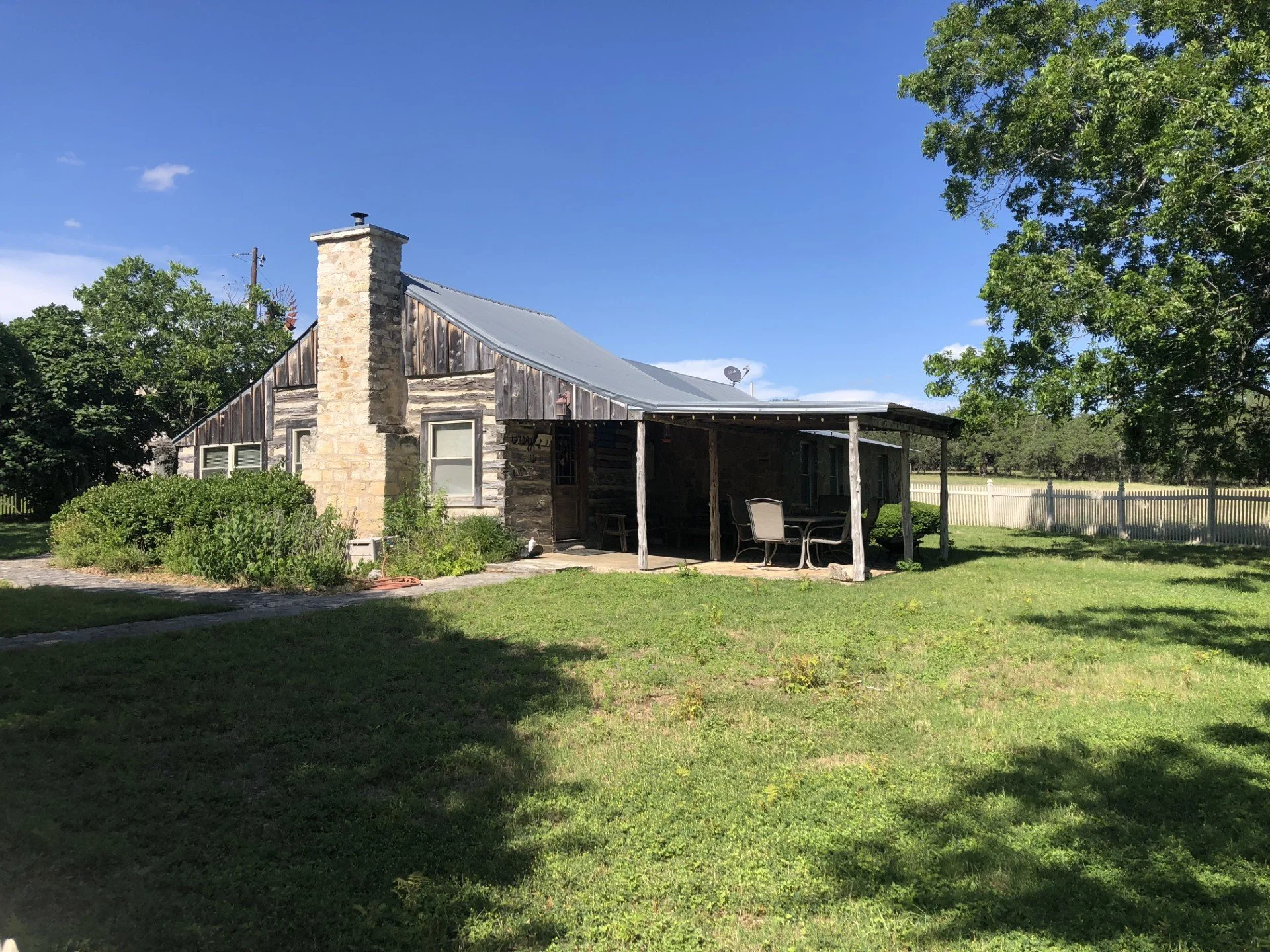 A rustic house with a stone chimney, wooden siding, and a metal roof. There is a wooden patio with outdoor furniture underneath a covered area. The yard is grassy with lush green trees and a white fence in the background.