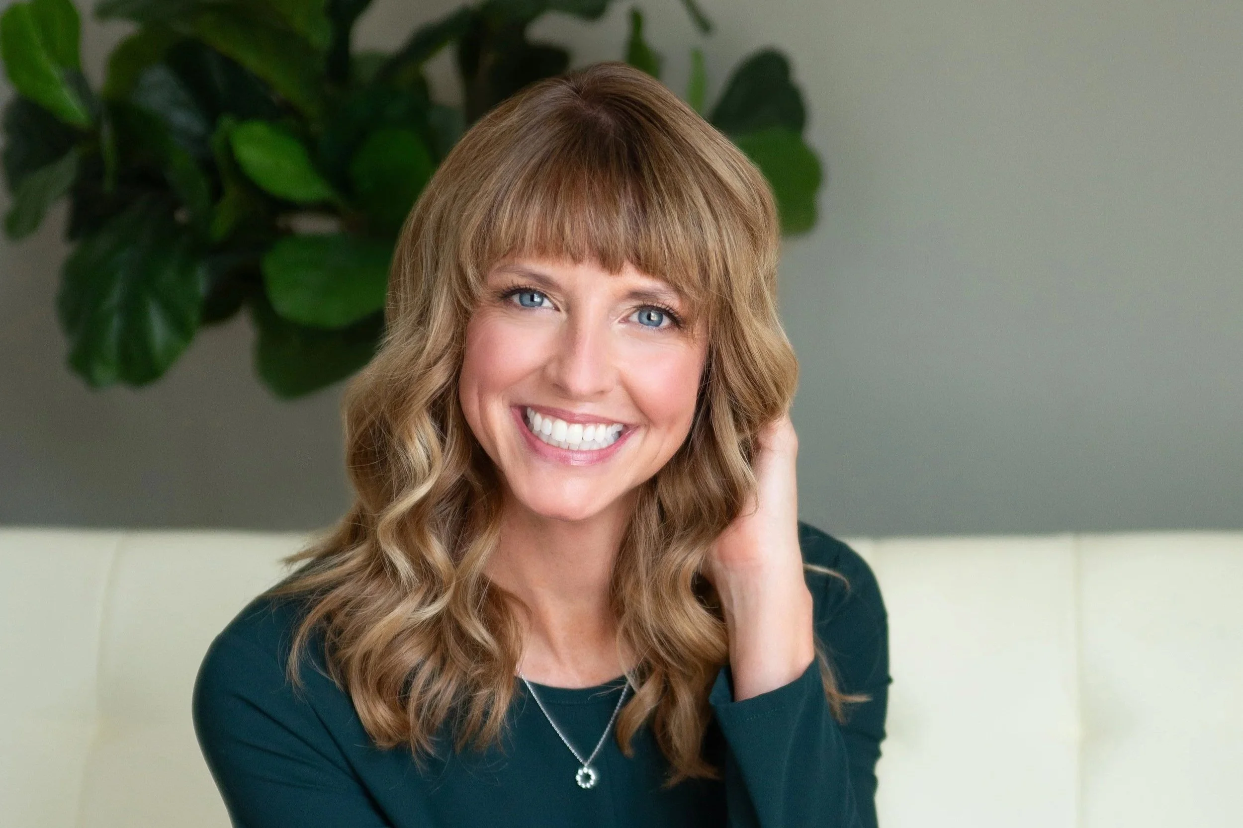 A woman with long, wavy blonde hair and blue eyes smiling at the camera in front of green plants.