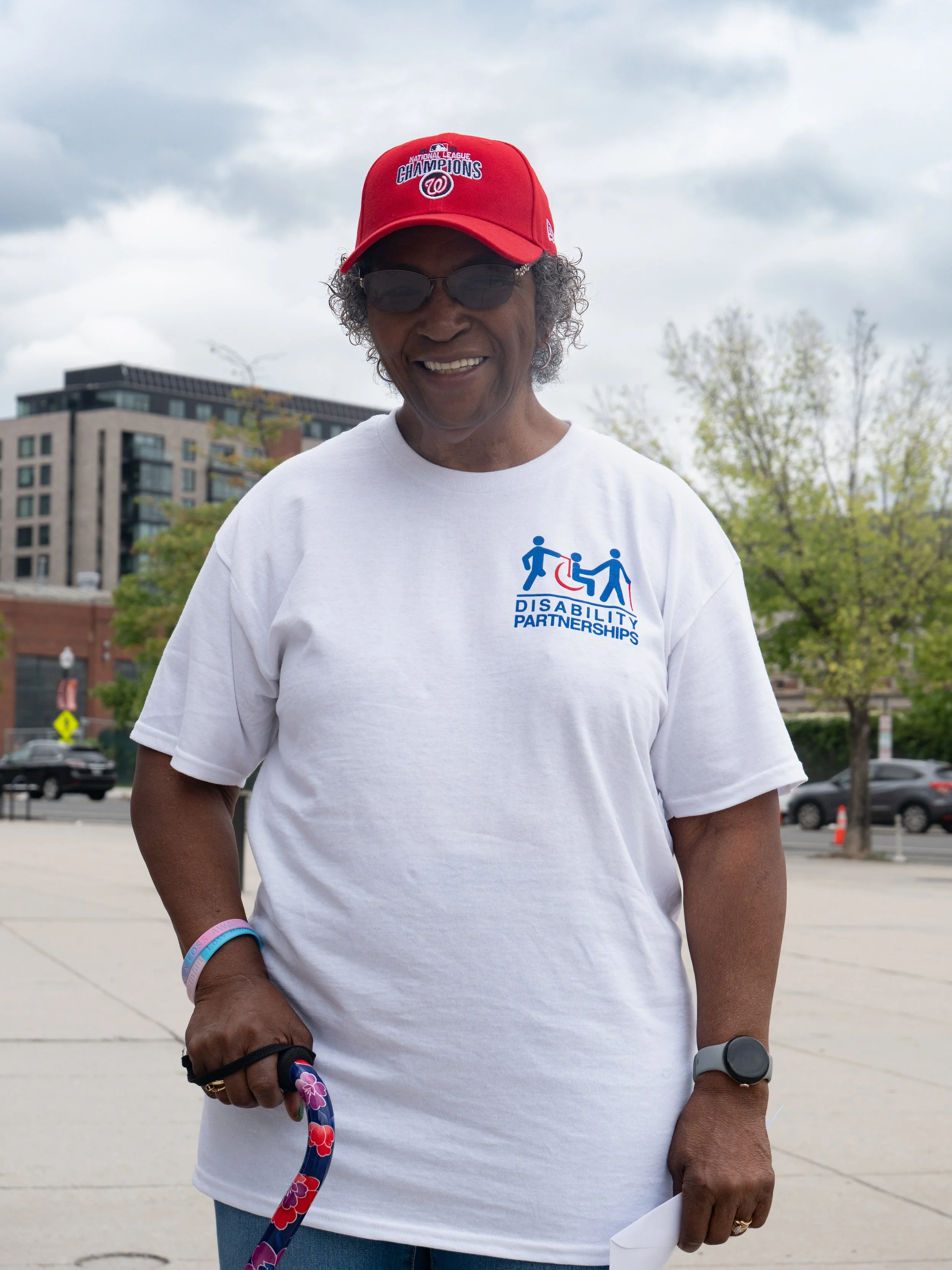 A smiling woman wearing a red Washington Nationals cap and a white Disability Partnerships T-shirt stands outdoors, holding a colorful floral cane.