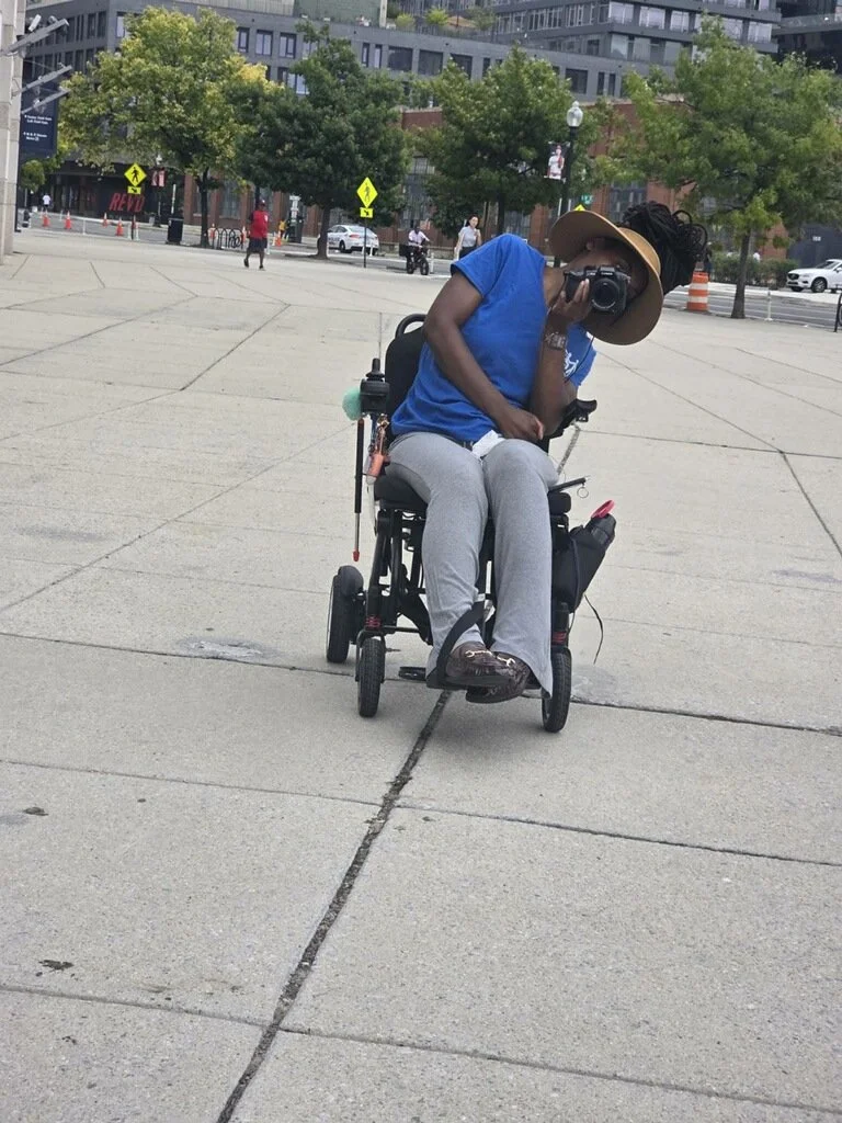 A photographer, named Tyree, using a power wheelchair takes a photo outdoors.