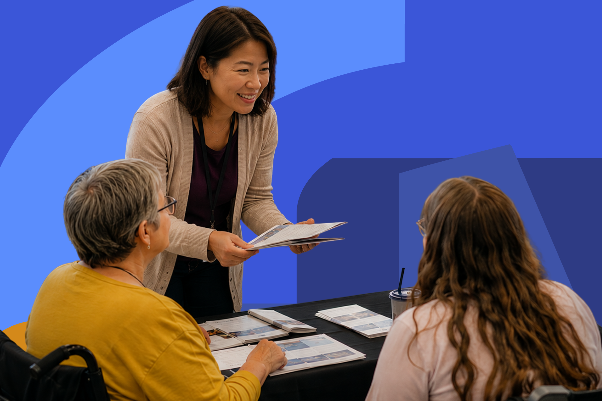 An Asian woman volunteer smiles as she welcomes participants at an inclusive community event. Around her, a diverse group of adults, including people with disabilities and wheelchair users, talk, laugh, and participate in activities together in a bri