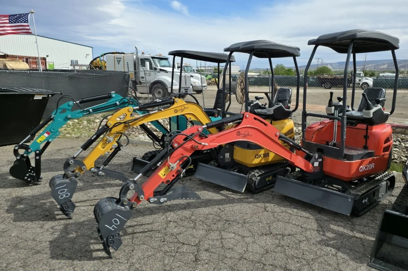 Three mini excavators in red, yellow, and teal colors with black tracks and canopies parked outdoors on a paved surface, with trucks and a chain-link fence in the background.