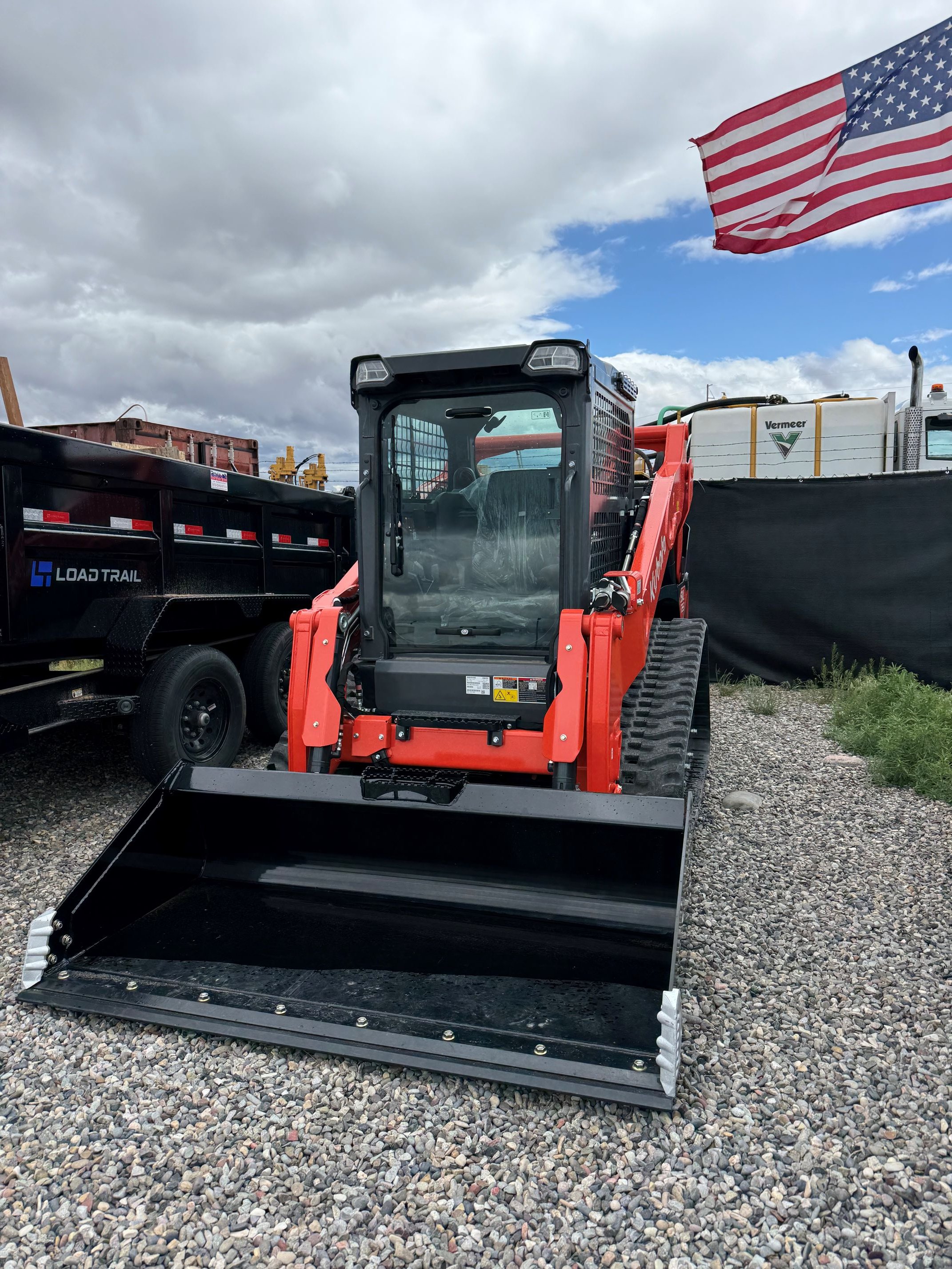 A construction vehicle with a black bucket, orange body, and enclosed cab, parked on gravel ground, with a black trailer and a white tank with green and black branding behind it, under a cloudy sky with an American flag flying.
