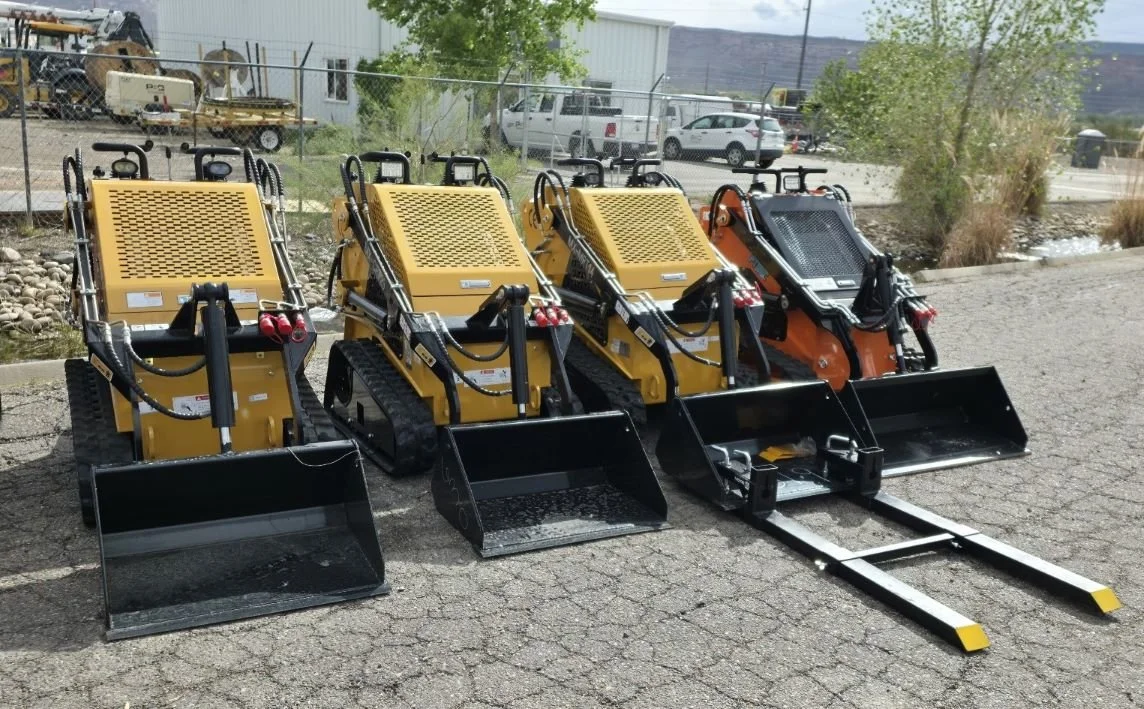 Four mini skid steer loaders with different attachments, lined up outdoors on a paved surface.