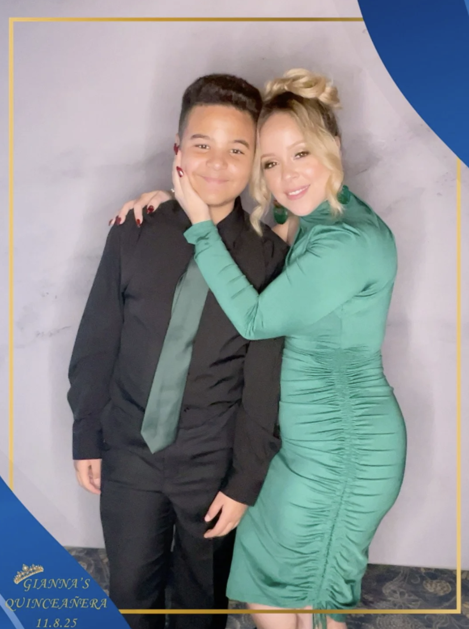 A woman in a green dress with earrings and blonde hair styled in an updo, hugging a young person with short hair, wearing a black outfit with a dark green tie, against a grey background.