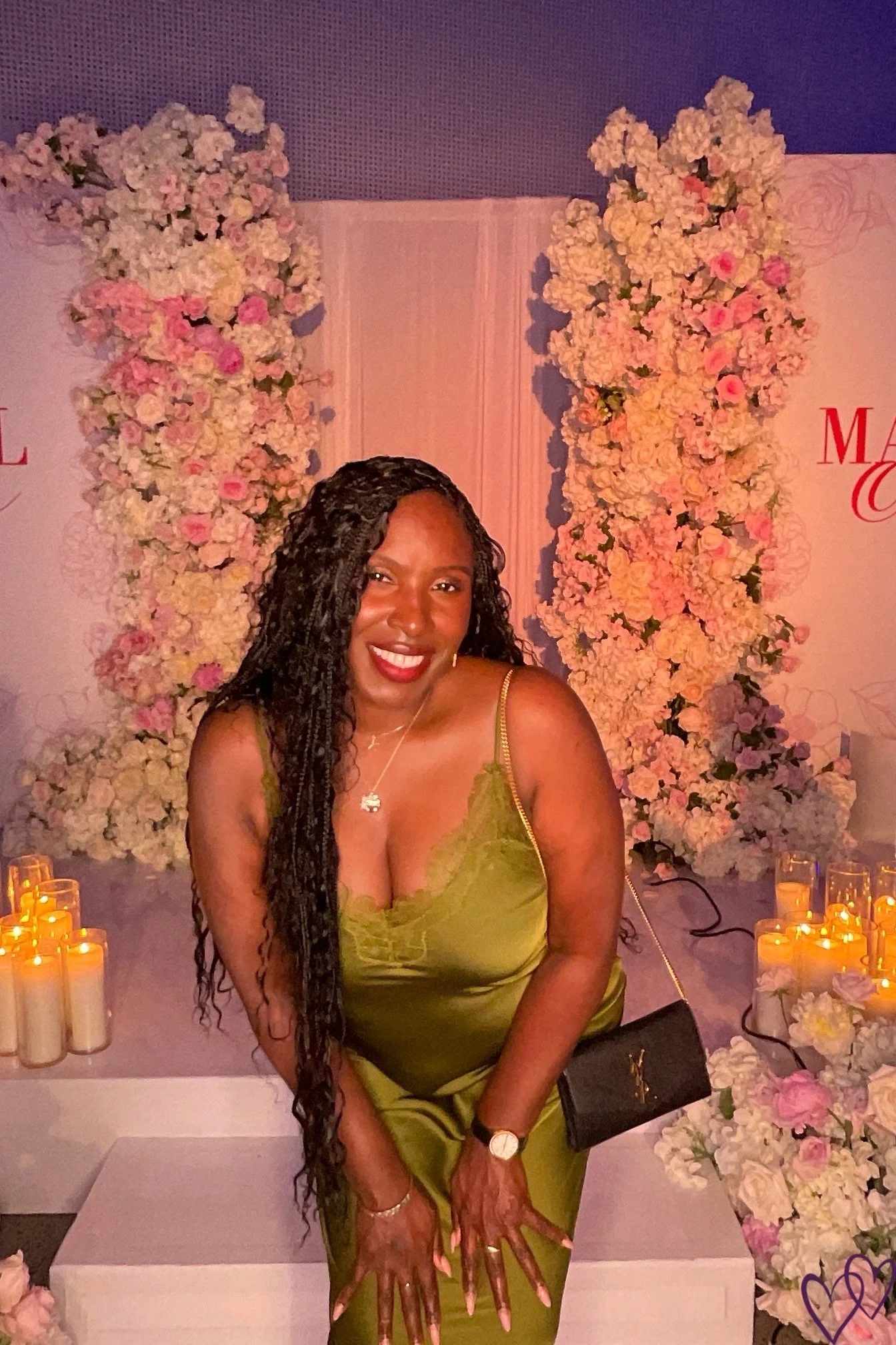 A woman with long, braided hair smiling at a celebration surrounded by pink and white flowers and lit candles.