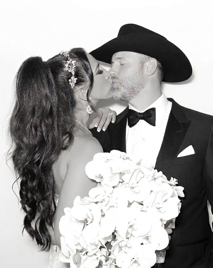 Black and white photo of a bride and groom sharing a kiss on their wedding day. The bride has long, wavy hair with a floral headpiece, and the groom wears a cowboy hat and tuxedo, holding a bouquet of white flowers.
