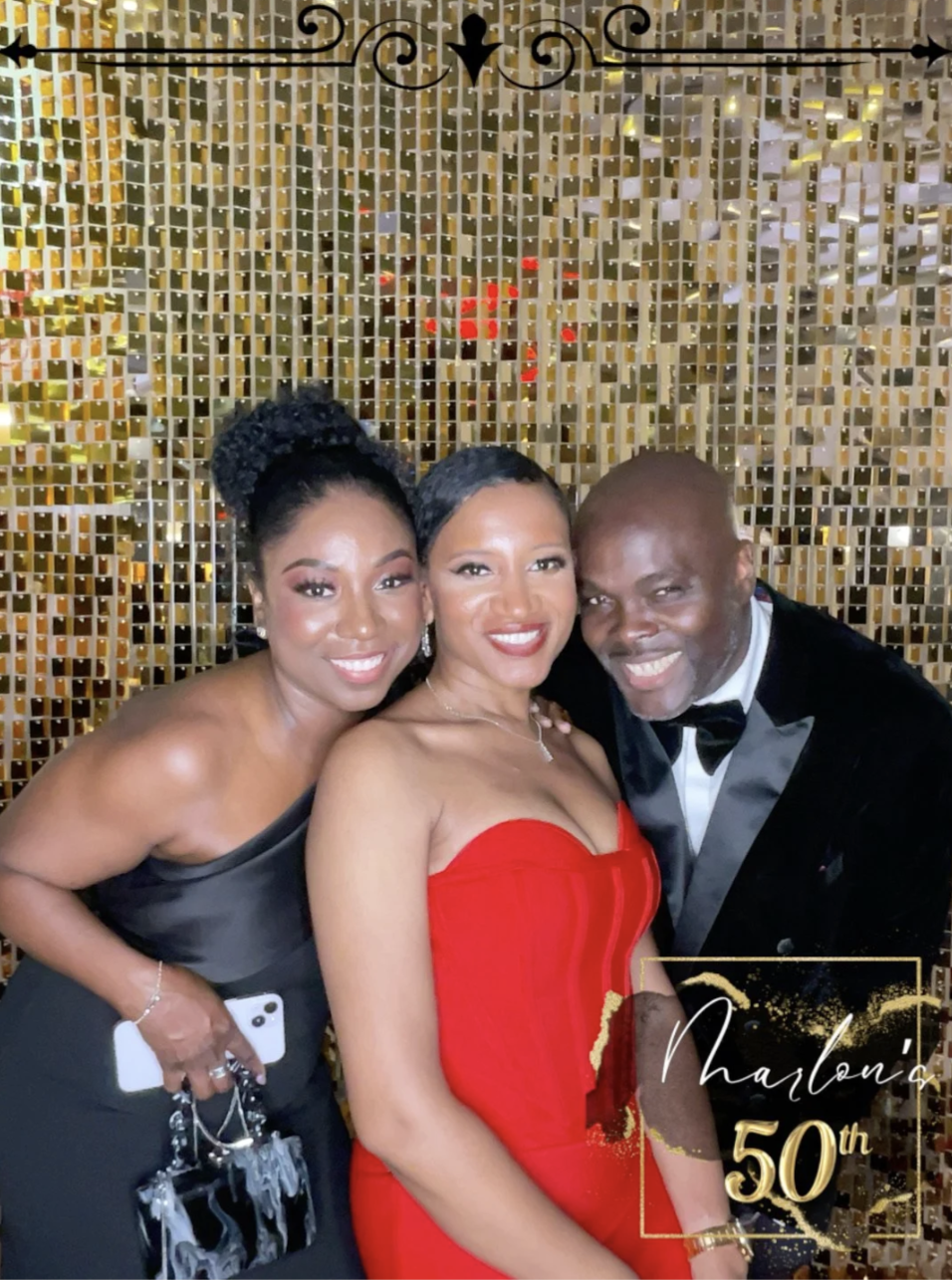 Three people dressed in formal attire at a celebration with a gold glitter backdrop. The woman on the left is in a black strapless gown, the woman in the middle is in a red strapless gown, and the man on the right is in a tuxedo. There is text on the
