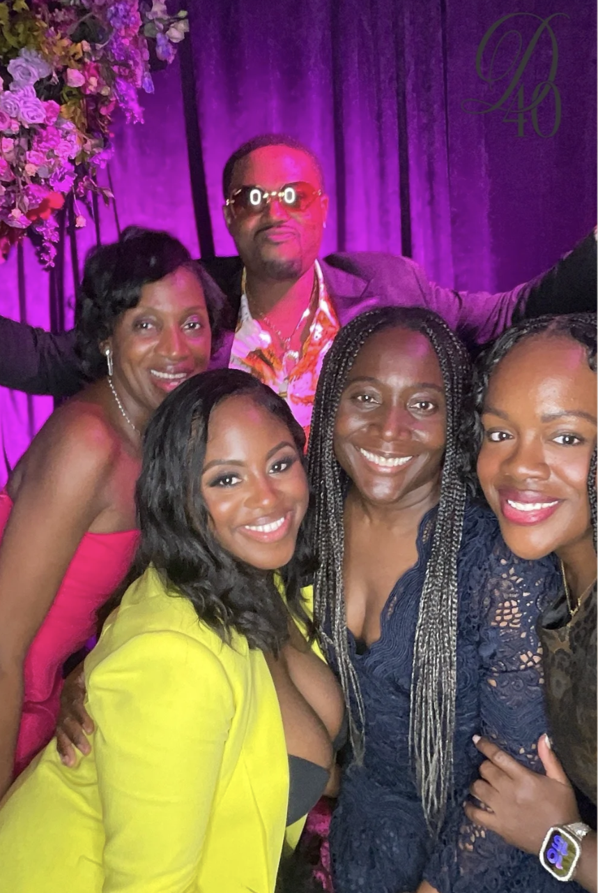 A group of five women and one man smiling at a celebration event with purple lighting, floral decorations, and a purple curtain background.