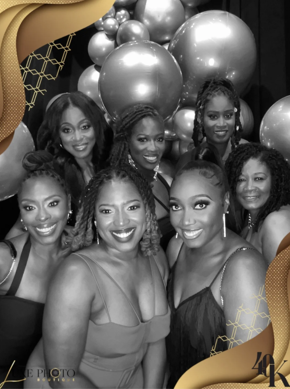 Group of seven women at a celebration, smiling and posing together in front of decorative balloons, with gold and black-themed framing.