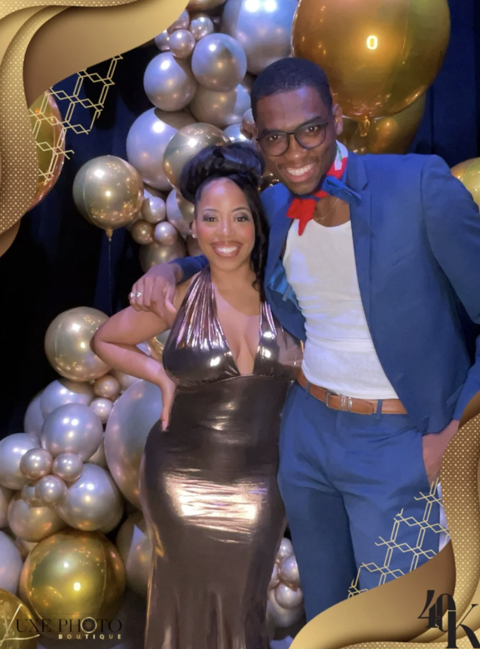 A smiling woman in a metallic gold dress and a smiling man in a blue suit at a party with gold and pearl balloons in the background.