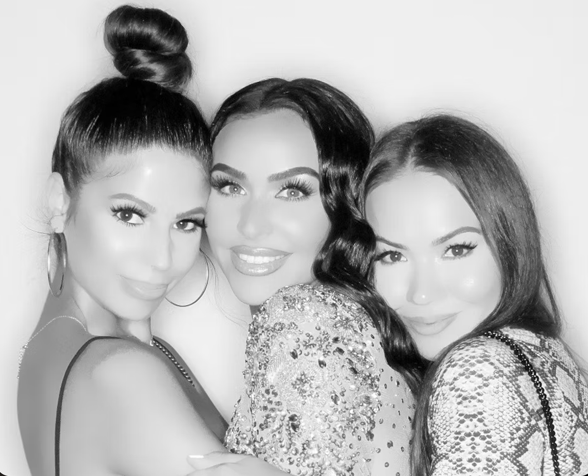 Three women in glamorous outfits, posing together in black-and-white. The woman in the middle has dark, wavy hair and wears a sequined top. The woman on the left has her hair styled in a sleek bun and wears large hoop earrings. The woman on the right