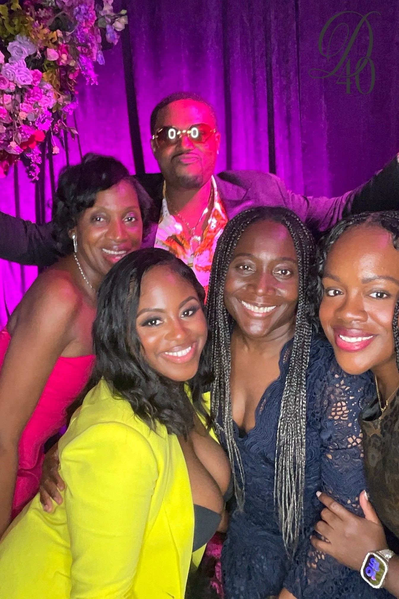 A group of five women and one man at a celebration event, smiling for the camera. The background is decorated with purple curtains, floral arrangements, and a logo that reads "D40." The women are dressed in colorful attire, and the man is wearing sun