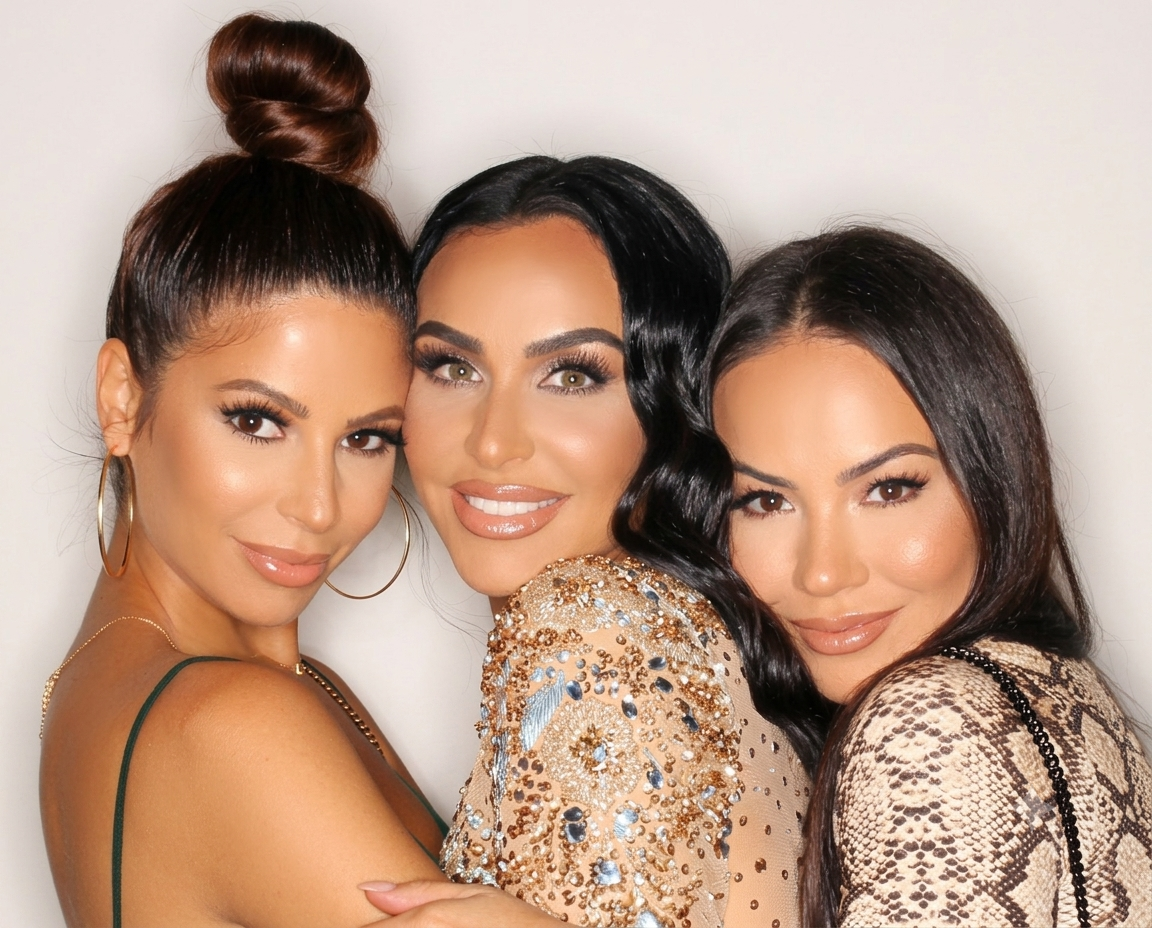 Three women smiling close together, one with a top knot hairstyle, one with black wavy hair, and one with long dark hair, posing for a photo indoors.
