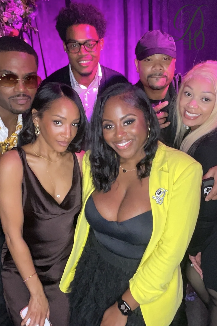 A group of six diverse people posing together at a celebration or event, with a purple background and flowers on the side. They are smiling and dressed in fashionable attire, with some wearing accessories like earrings and necklaces.