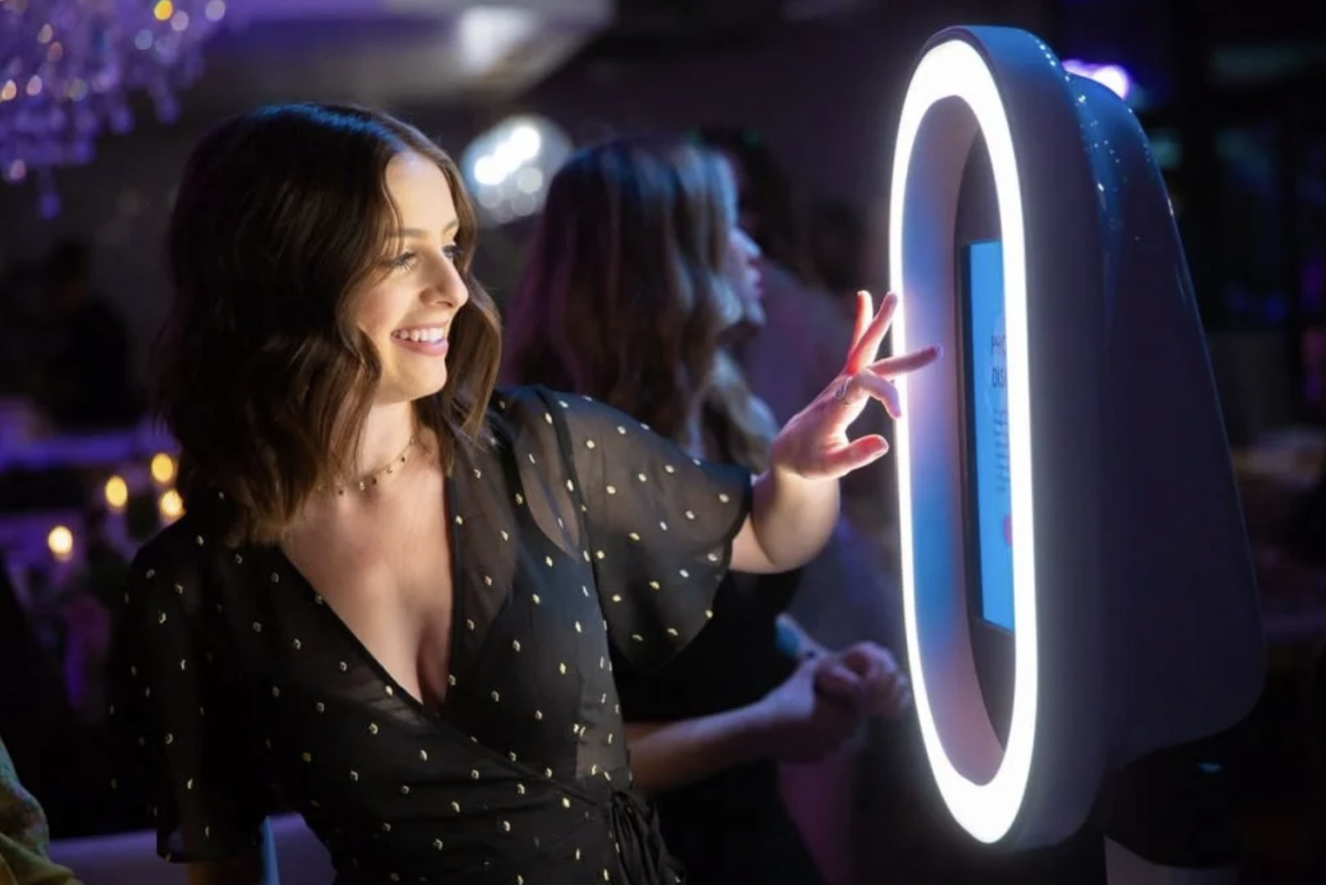 A woman with wavy brown hair smiling and pressing a button on an arcade game where she is making a peace sign with her fingers. She is in a dark room with colorful lights in the background.