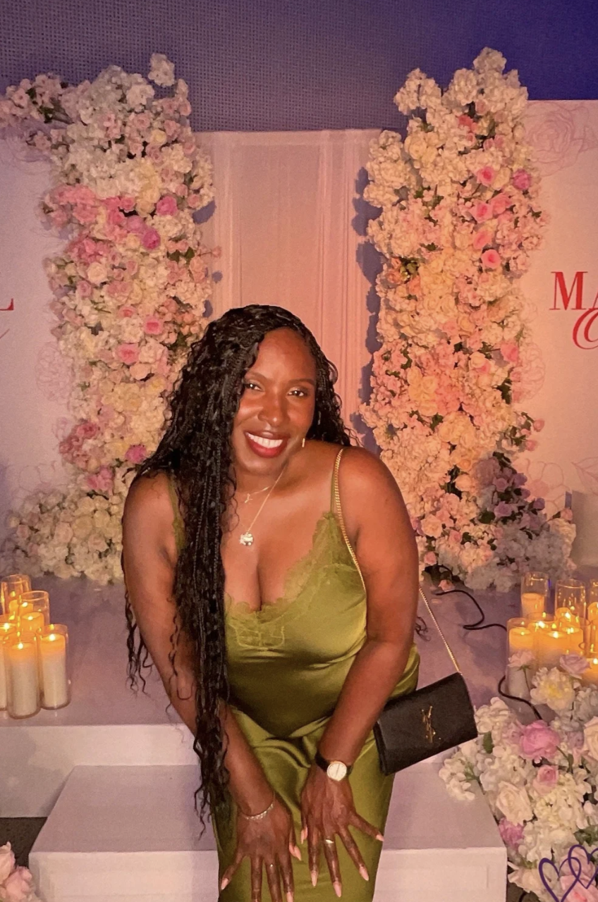 A woman in a green satin dress with long black curly hair, smiling at a floral event with pink and white flowers and candles in the background.