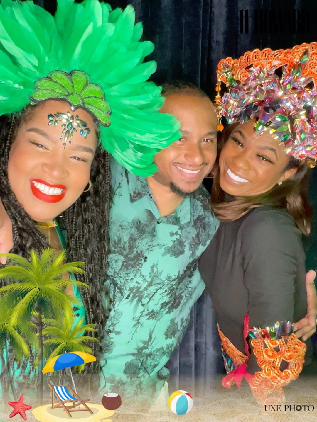 Three people smiling, wearing colorful headdresses; two women in decorative costumes, one man in Hawaiian shirt, celebrating at a festive event.