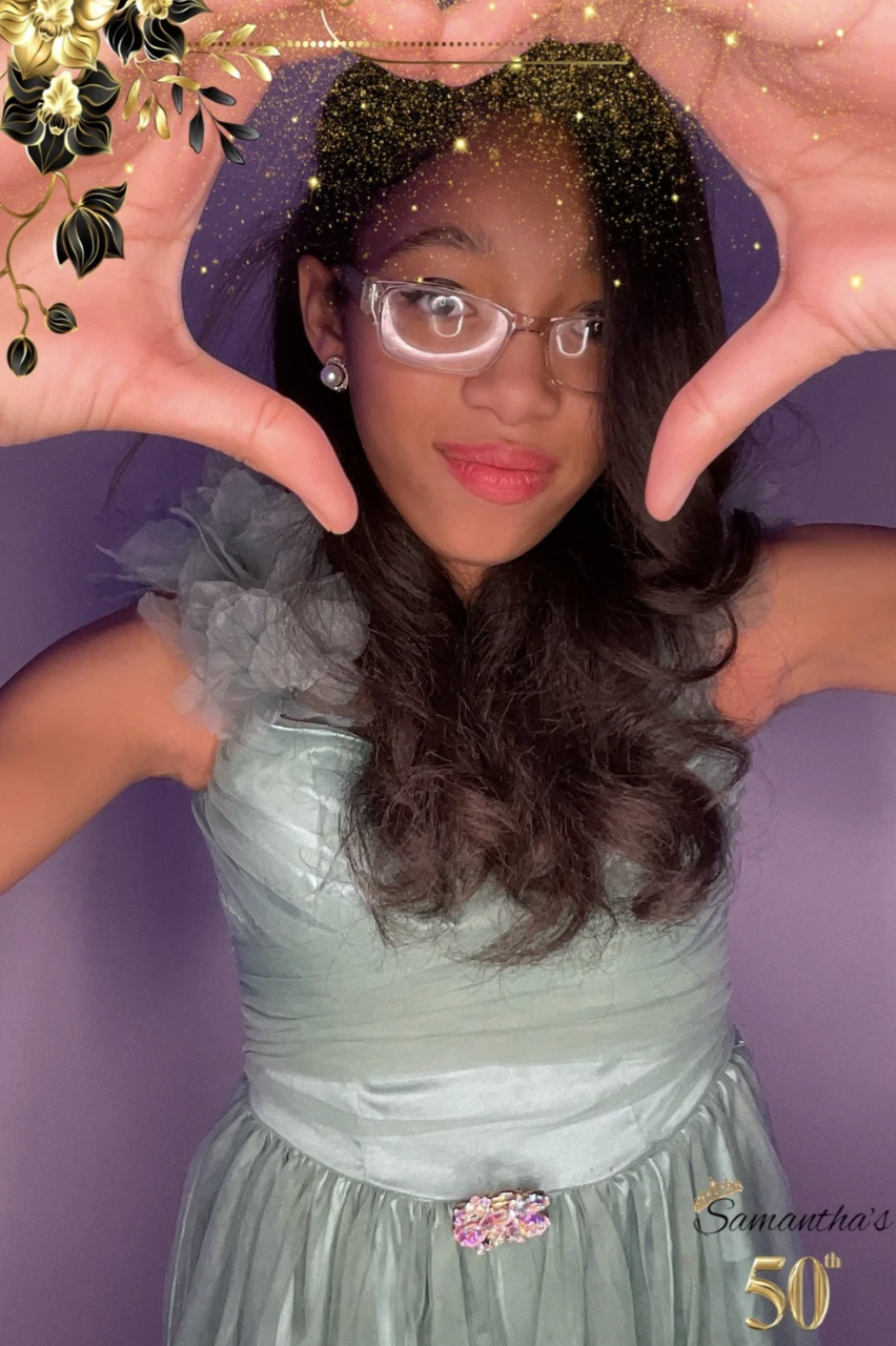 A woman wearing a light-colored dress with ruffled shoulders and a decorative belt, posing with her hands framing her face, and smiling. She has long, dark hair, wears glasses and earrings, and is standing against a purple background with a floral an