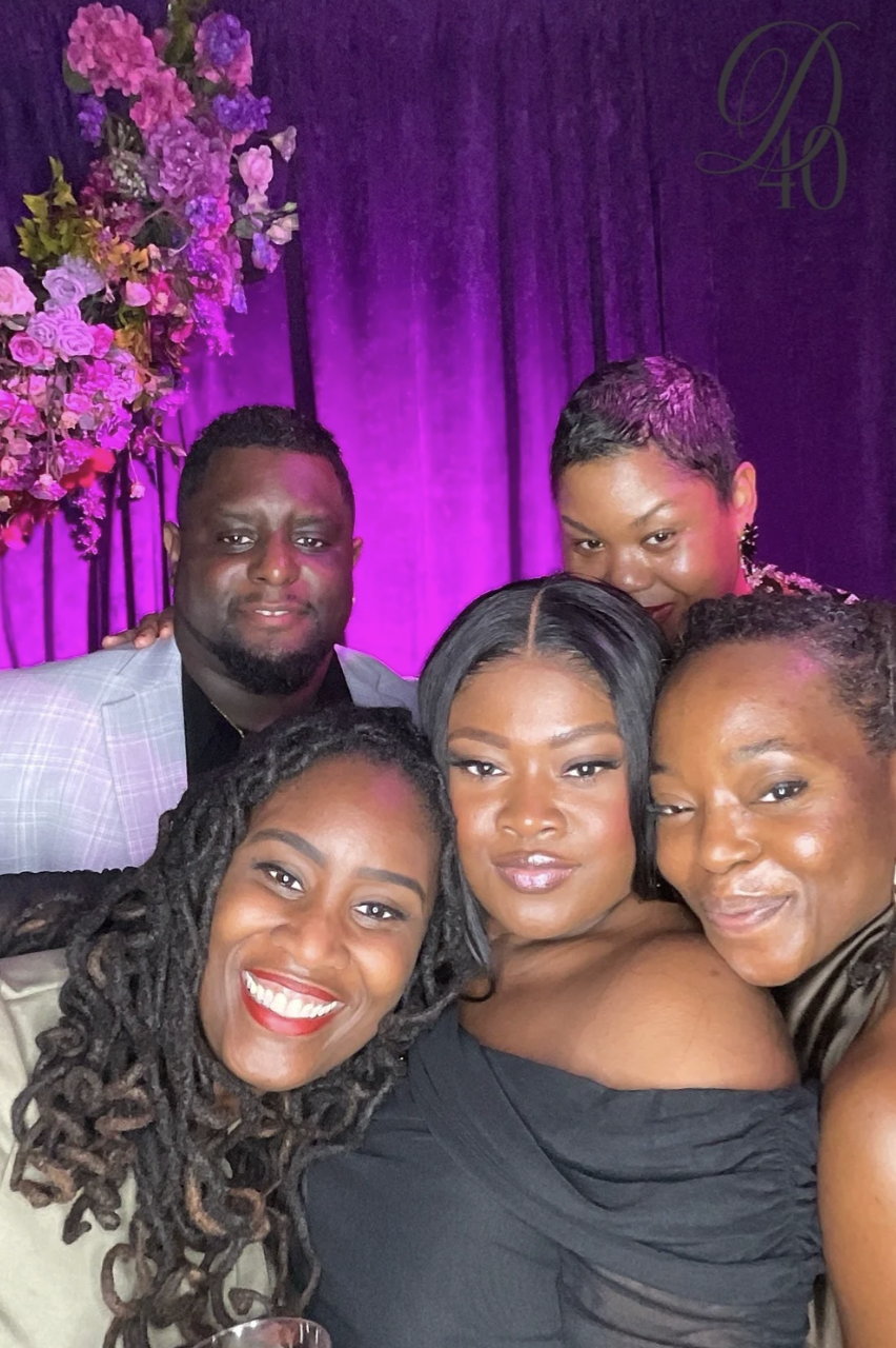 Group of five women and one man taking a selfie at a celebration, with purple lighting and floral decorations in the background.