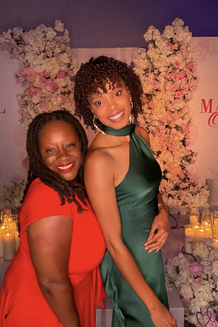 Two women standing together at an event with floral decorations and candles in the background. One woman is wearing a red dress and the other a dark green dress.