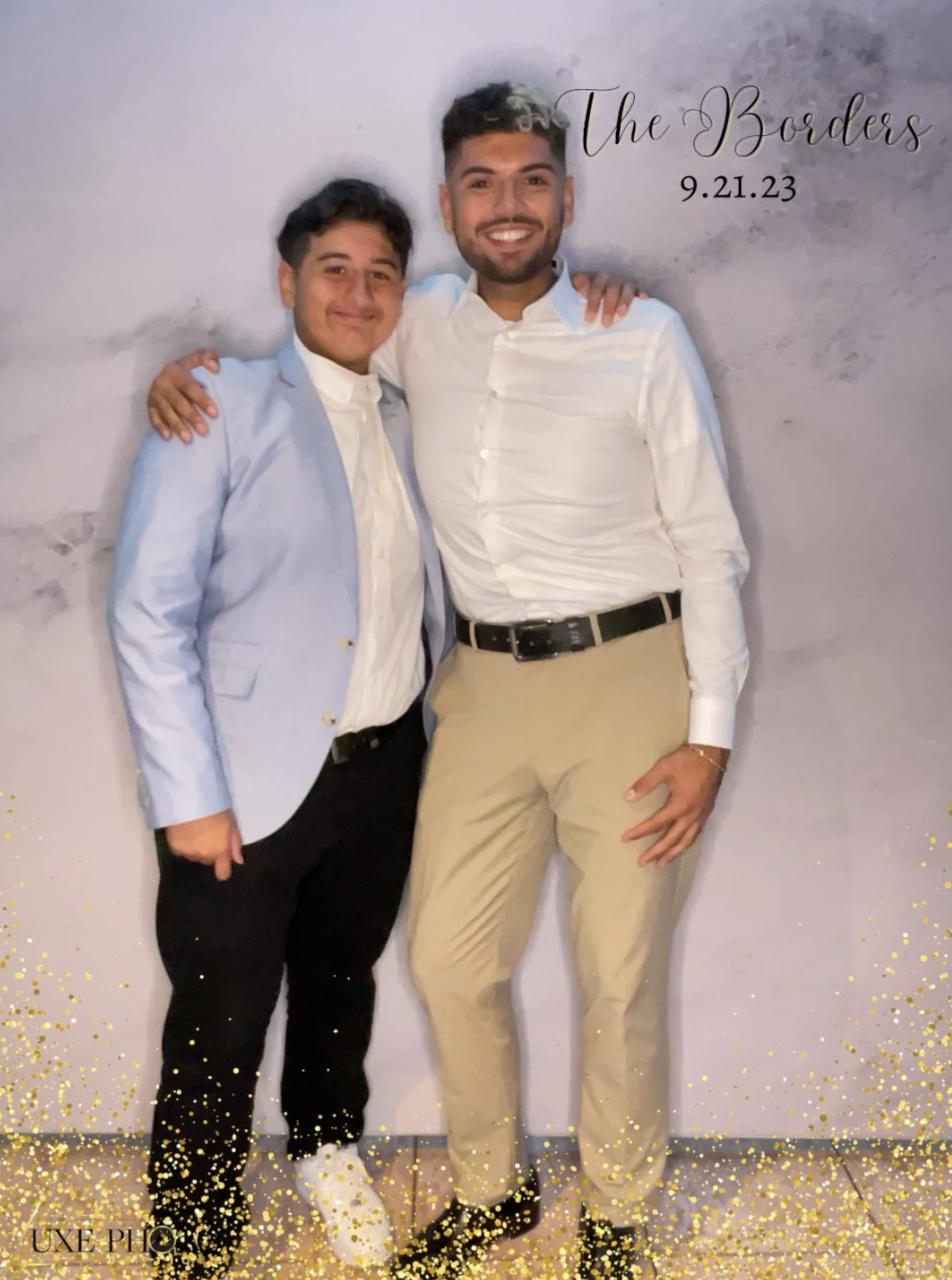 Two young men with dark hair smiling and standing close together, one wearing a light blue blazer and the other a white dress shirt and beige pants, posing in front of a backdrop that reads "The Borders 9.21.23" with gold glitter accents at the botto
