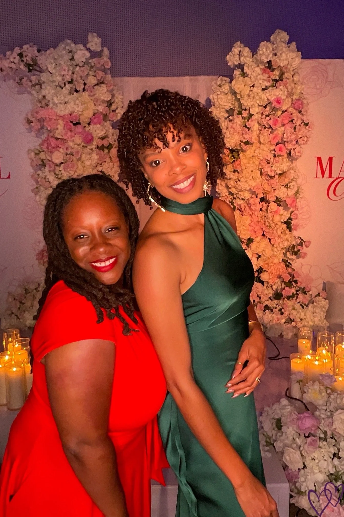 Two women smiling, one in a red dress and the other in a green dress, standing in front of a floral backdrop with pink and white flowers and candles.