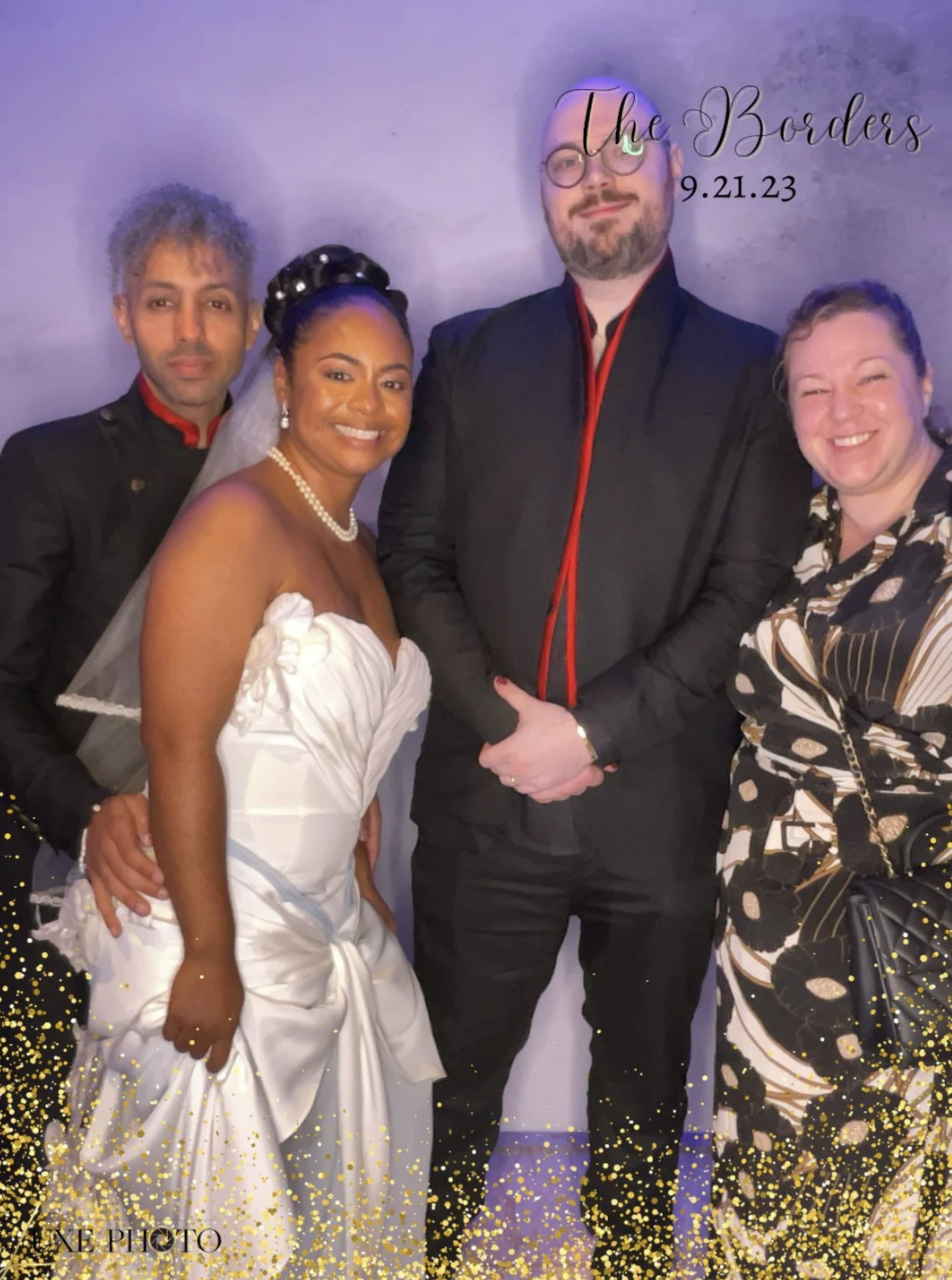 Four people at a wedding reception, with a bride in a white wedding gown and pearl necklace, a groom in a black suit with red accents, and two guests, all smiling and posing together against a purple backdrop with the text 'The Bouders' and date '9.2