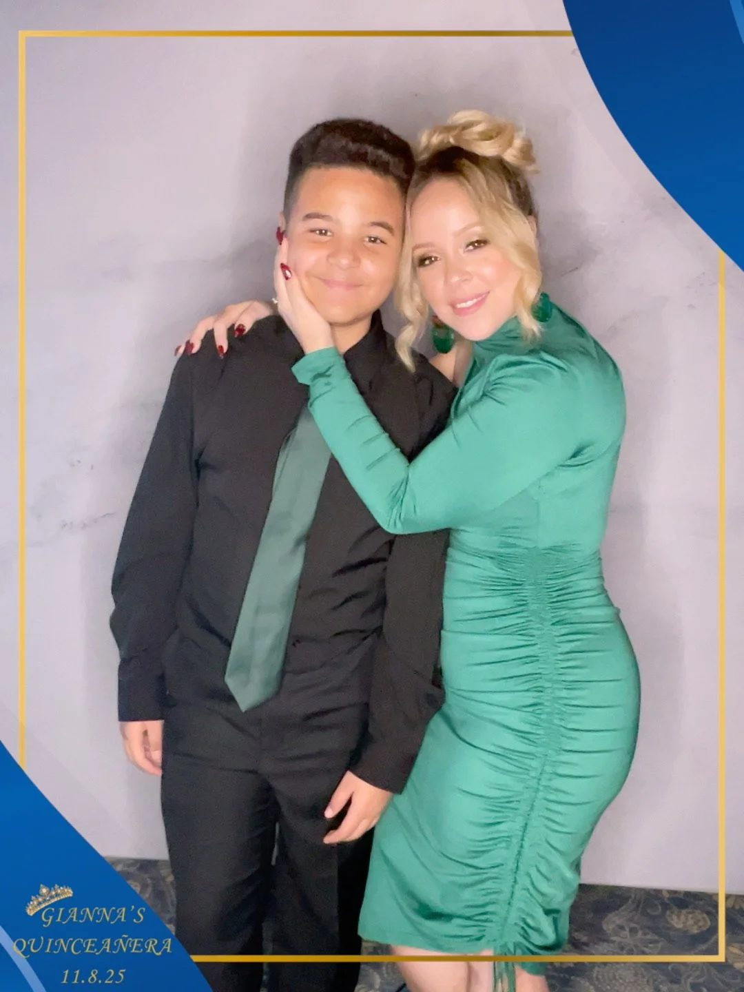A woman and a young boy posing together, smiling, with the woman holding the boy's face. The woman is wearing a tight, ruched green dress, and the boy is dressed in a black shirt with a green tie. The photo appears to have been taken at a celebratory