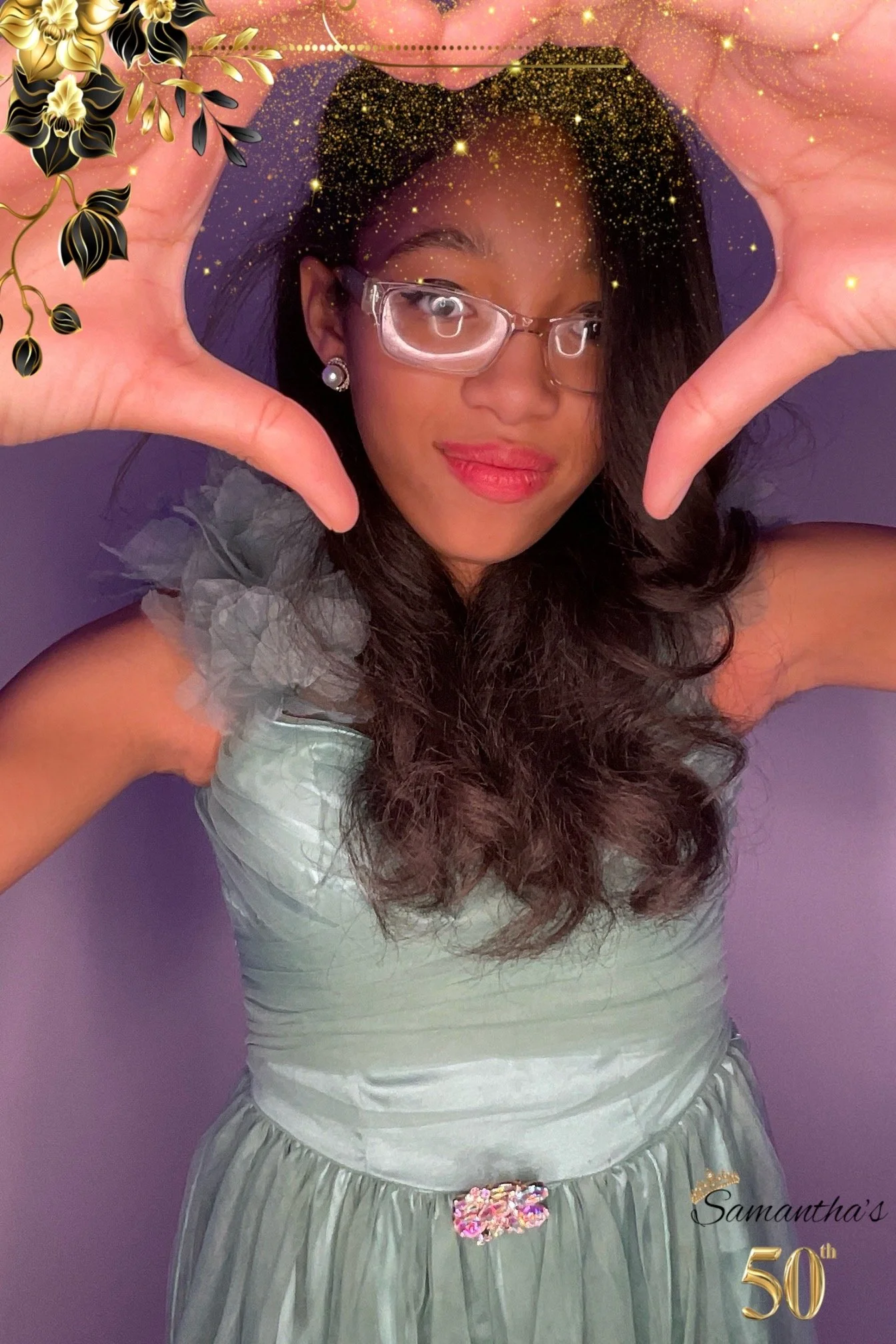A woman with long dark hair wearing glasses, pearl earrings, and a silver dress, holding her hands in a heart shape around her face, standing against a purple background with golden sparkles and floral decorations. There is a decorative text that rea