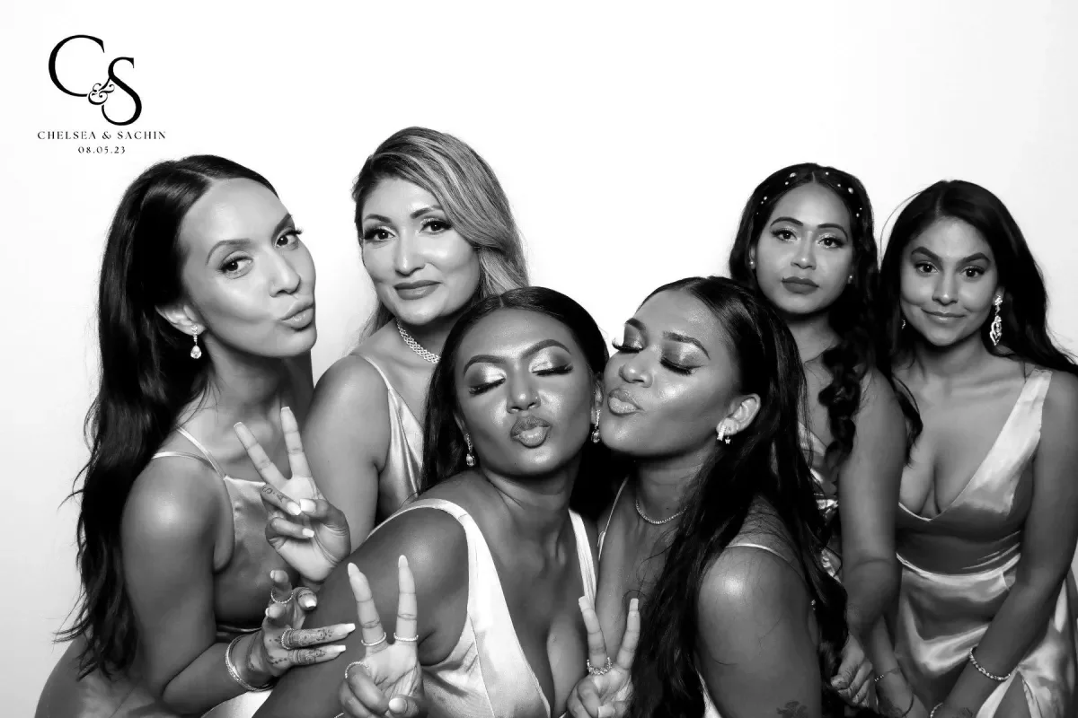 Black and white photo of six women posing together at a celebration, some making peace signs and kissing, wearing elegant dresses, with a logo and date in the top left corner.