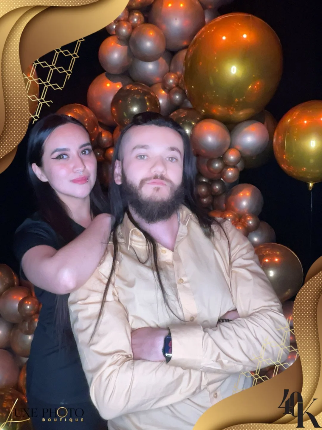 A man with long hair and a beard in a beige shirt stands with crossed arms, while a woman with dark hair and makeup stands behind him, touching his shoulder. They pose in front of a backdrop of gold and bronze balloons of various sizes, with decorati