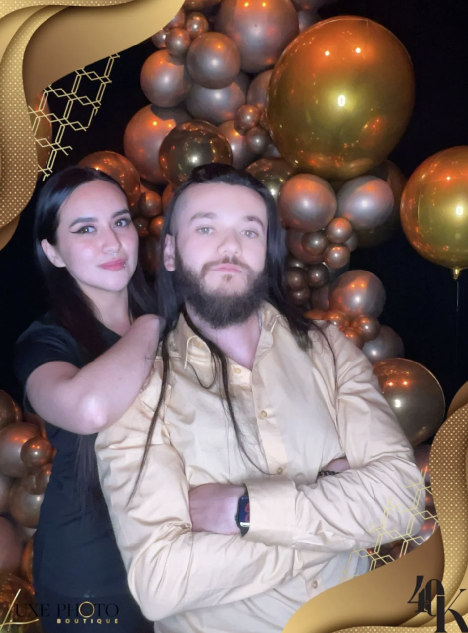 A man with long dark hair and a beard, wearing a beige shirt, stands with his arms crossed, posing in front of a backdrop of gold and bronze balloons and decorations, with a woman standing behind him to the left, touching his shoulder, both smiling a