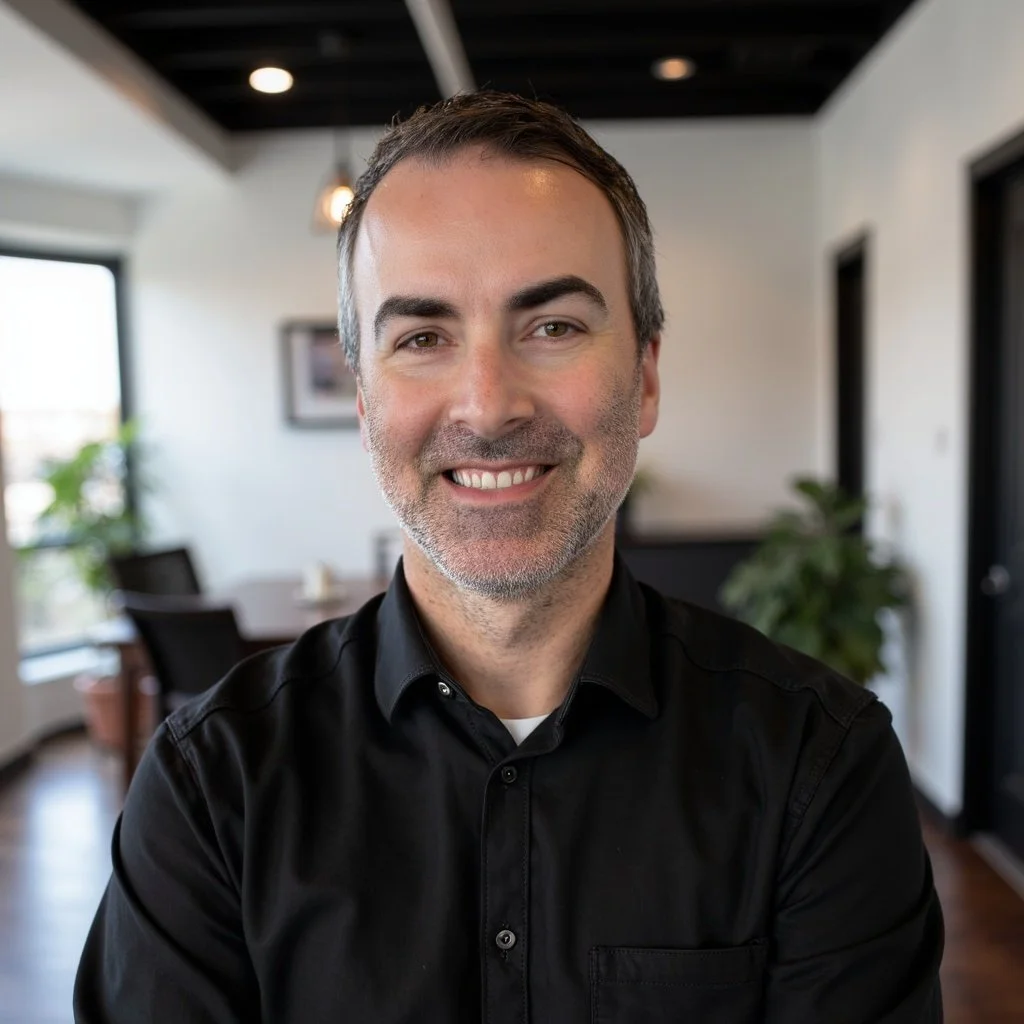 A smiling man with short dark hair and a beard, wearing a black shirt, sitting in a modern office or conference room with large windows, plants, and artwork on the walls.