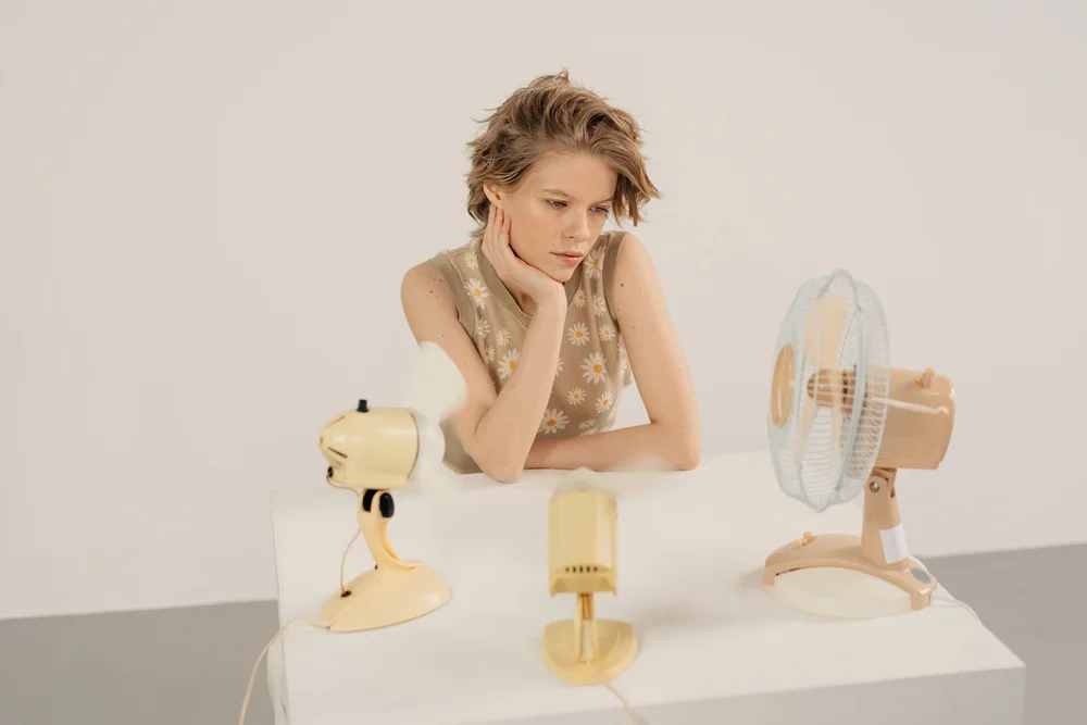 A woman seated near a small fan looking thoughtful