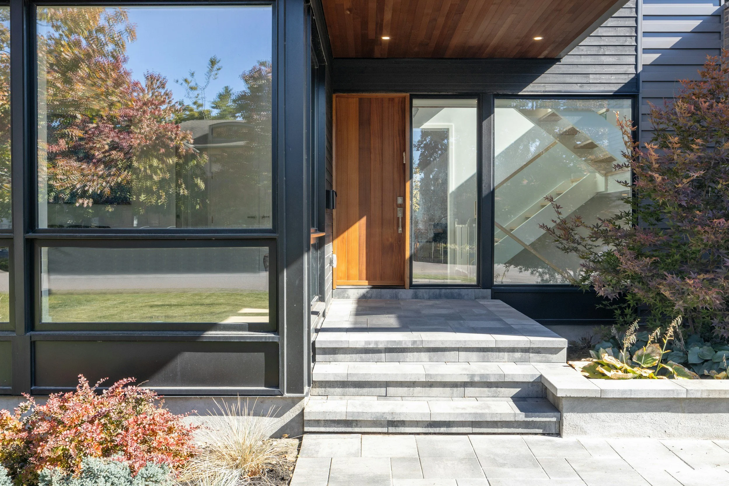 Contemporary house entrance with large glass windows, wooden door, and stone steps, surrounded by garden plants and trees.