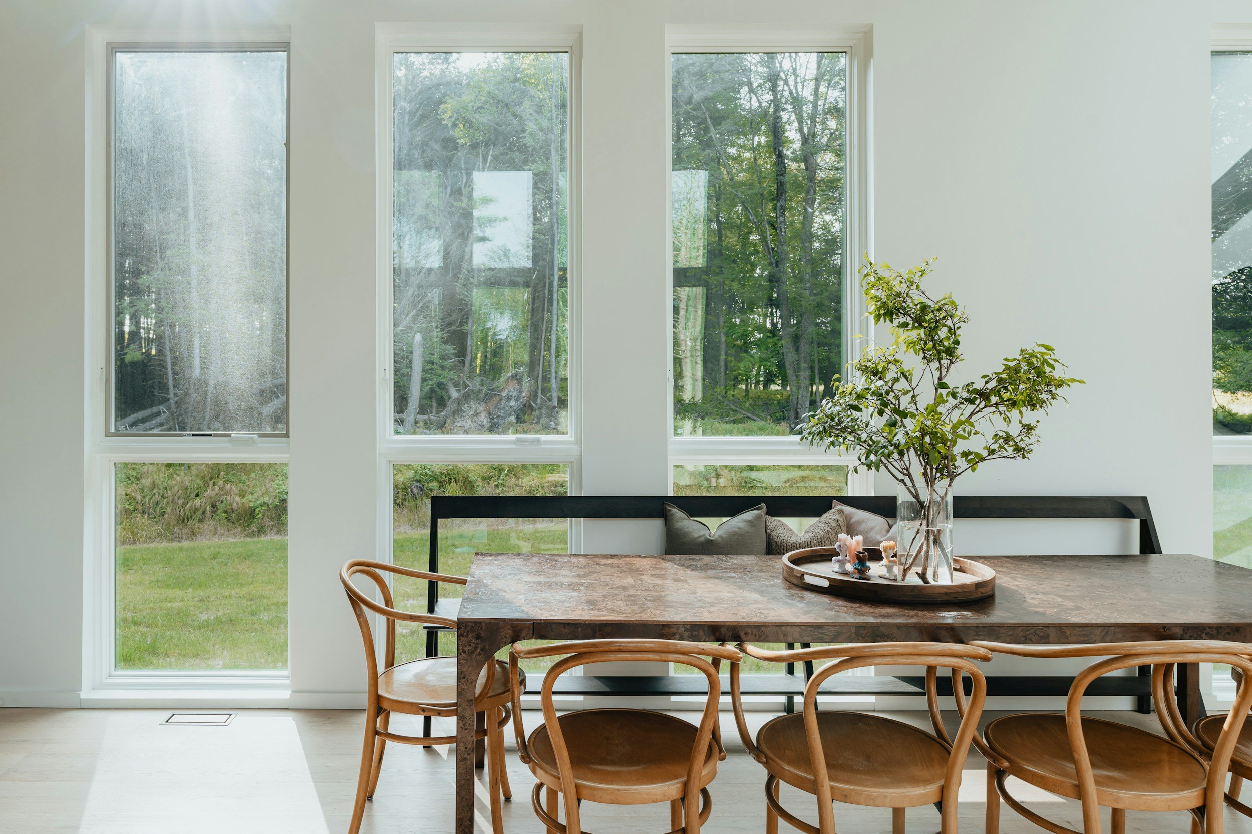Interior of a modern dining area with large windows showing a green garden outside, wooden chairs around a table with a decorative centerpiece, and a black bench with cushions.