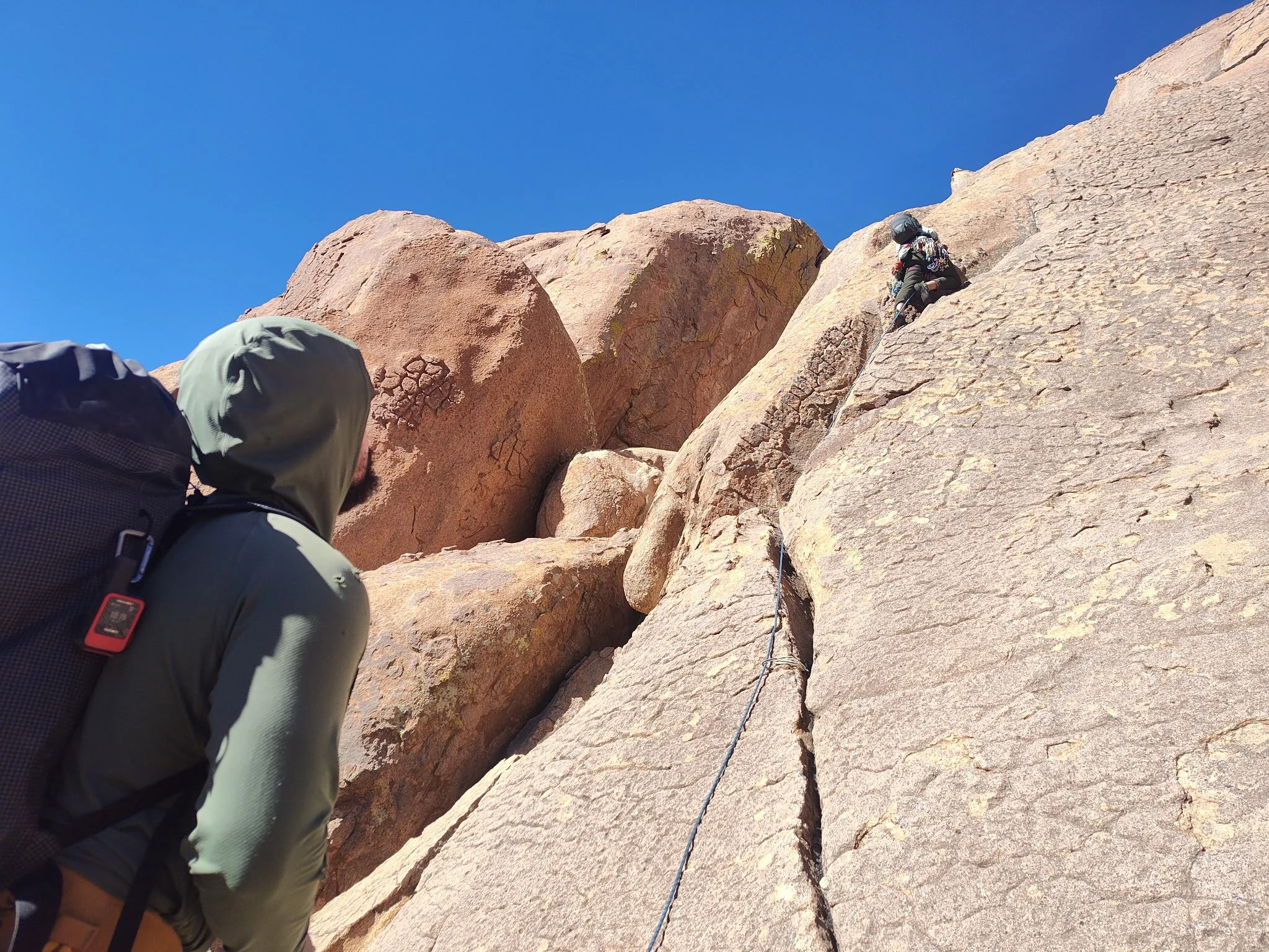 A person wearing a helmet and harness rock climbing on a steep, reddish-brown granite cliff under a clear blue sky.