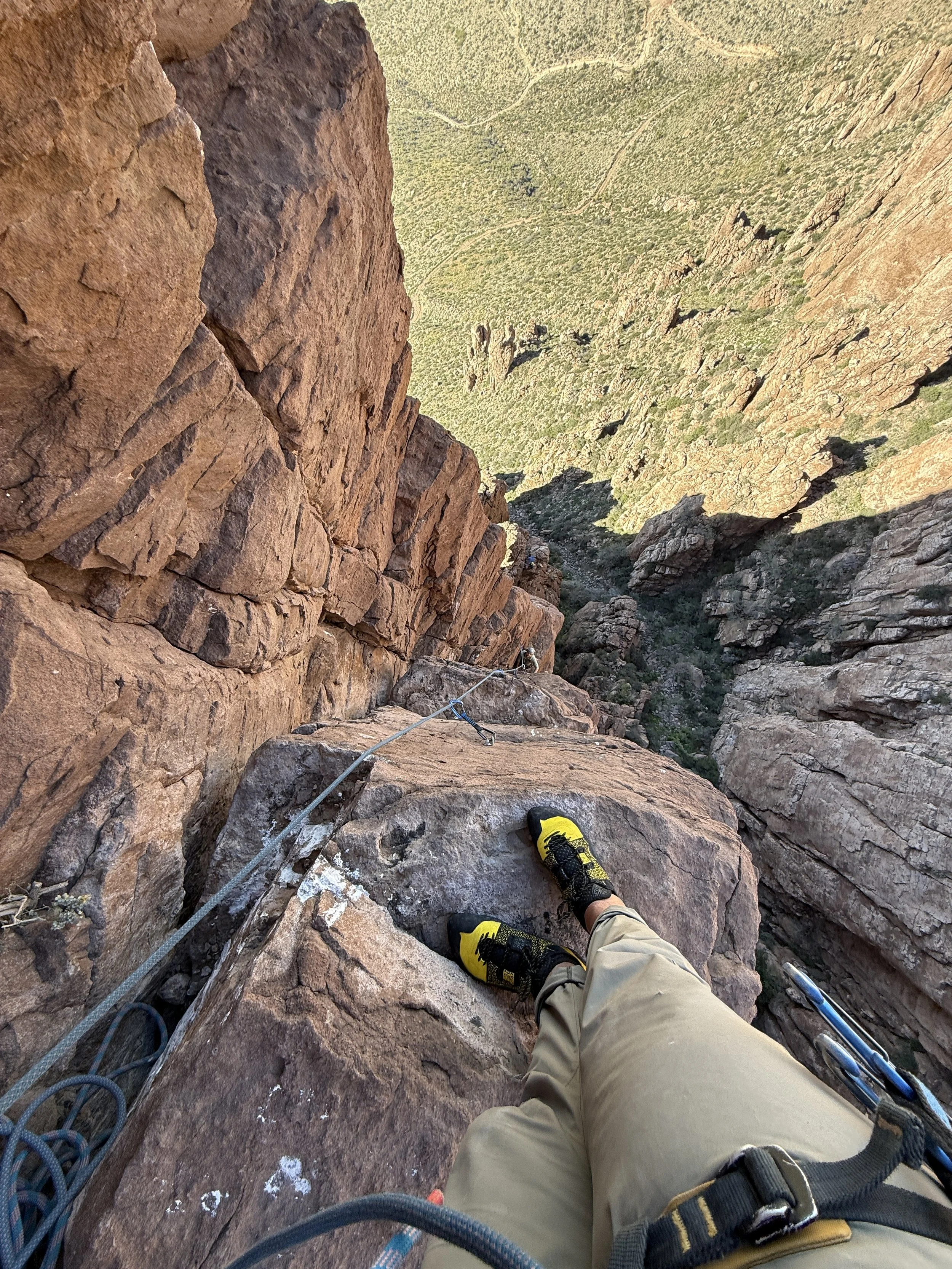 Looking down from a high cliff, a person wearing yellow and black climbing shoes and beige pants is secured with climbing gear and rope, with a vast green mountain landscape below.