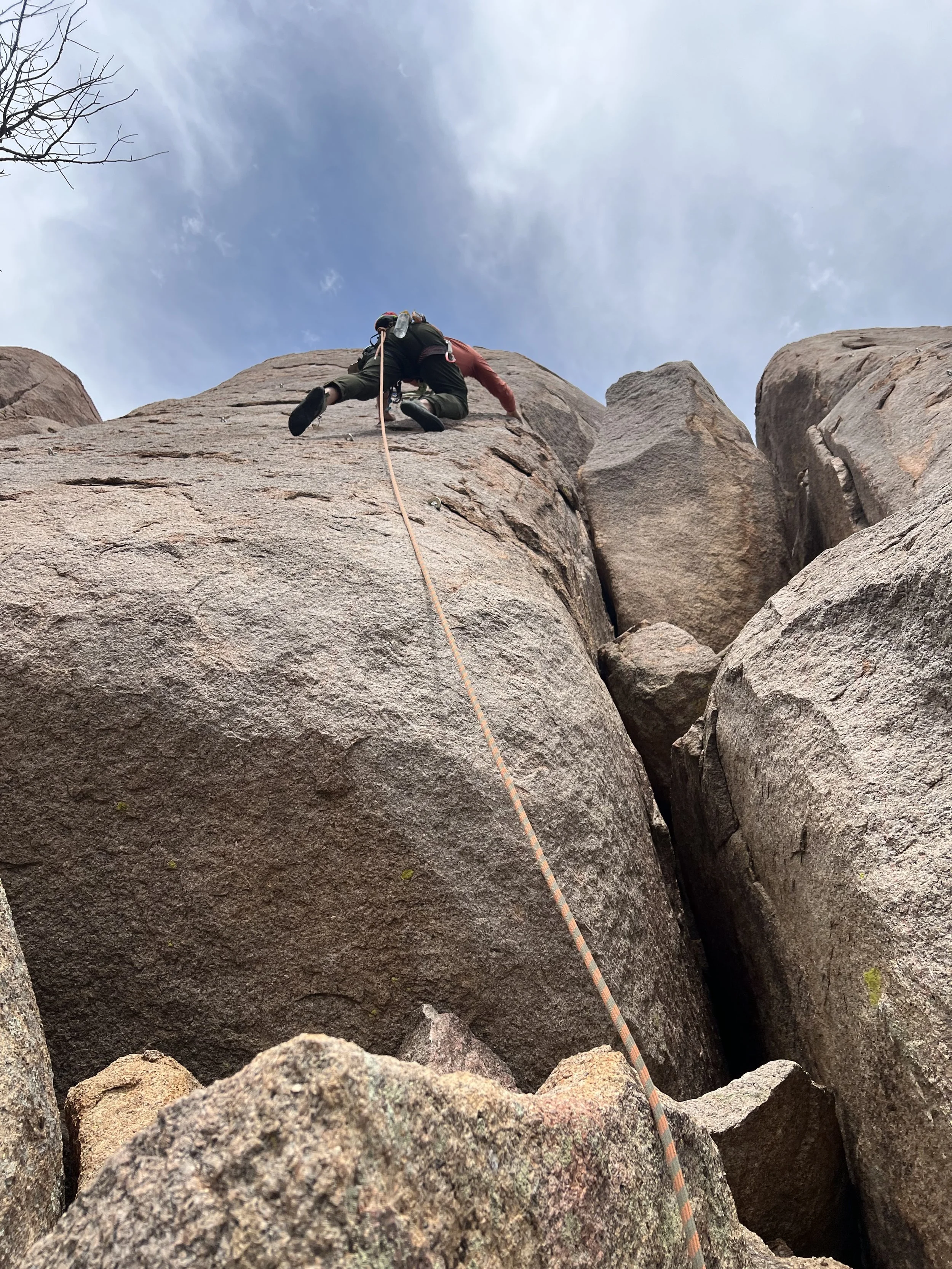 A person wearing climbing gear and helmet ascending a large rock face, with climbing rope attached, under a cloudy sky.
