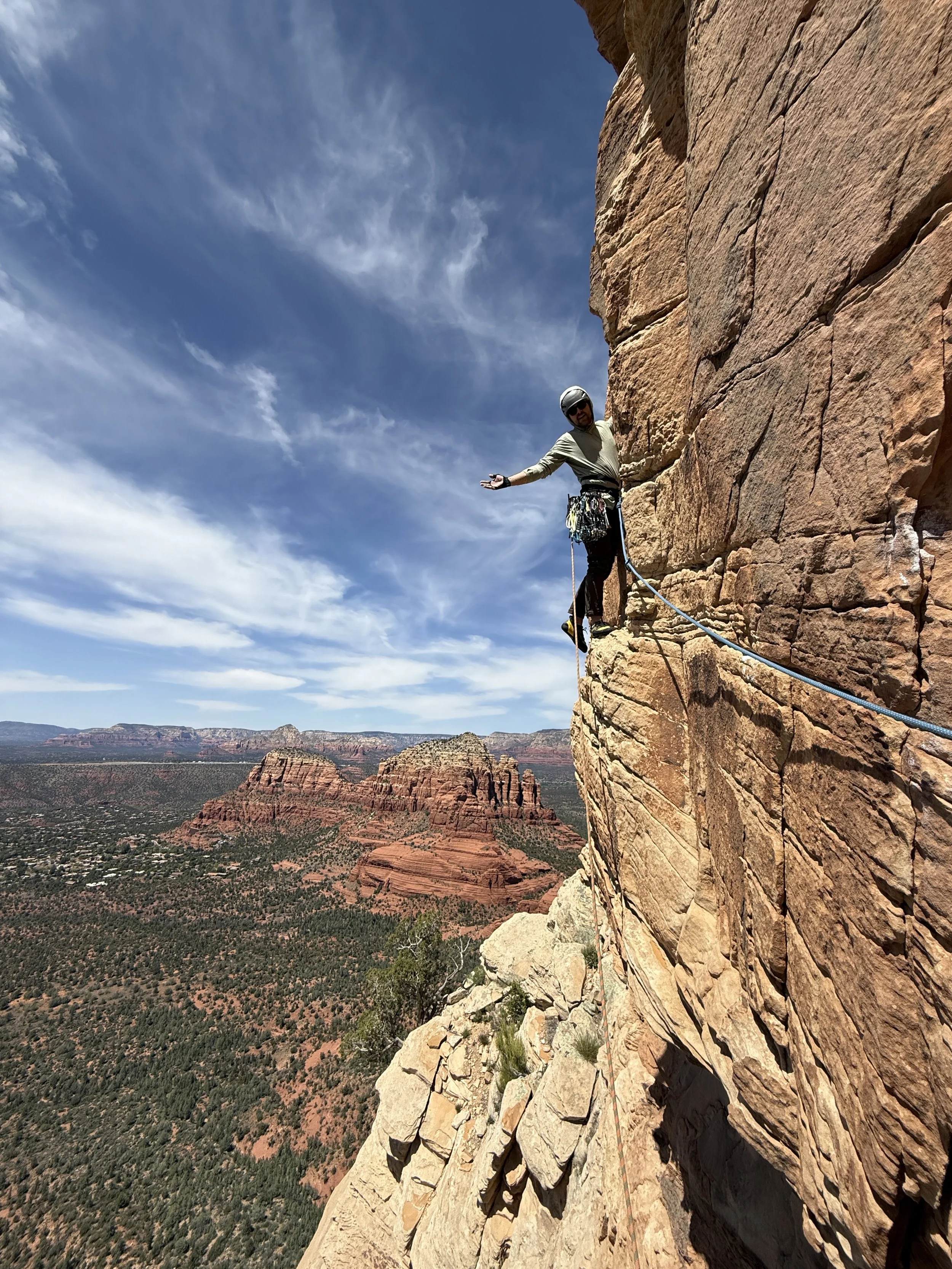 A man rock climbing on a sheer red cliff face in a desert landscape with red rock formations and pine trees, under a blue sky with wispy clouds.