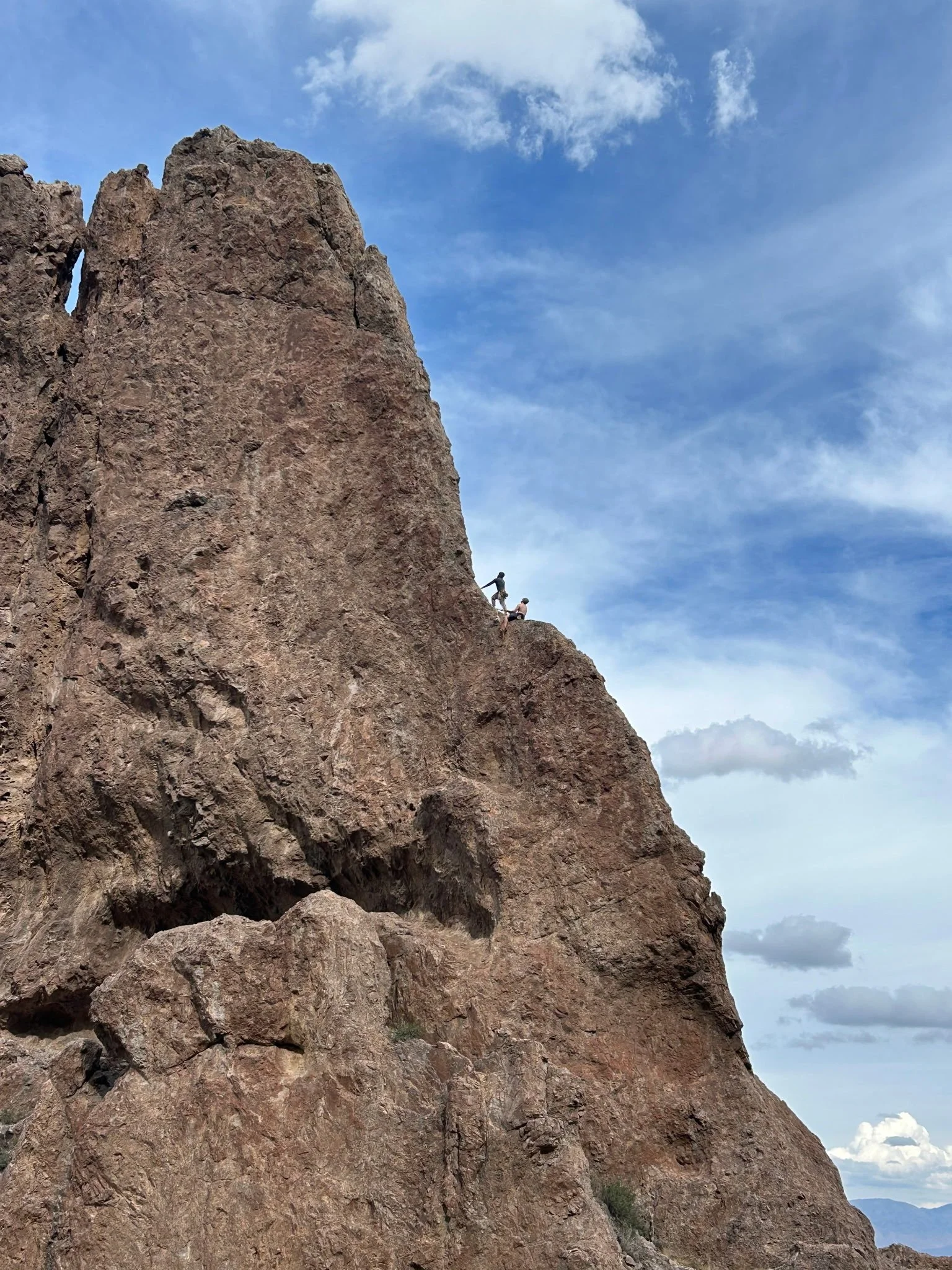 Two people rock climbing on a large, brown rocky mountain with a blue sky and scattered clouds in the background.
