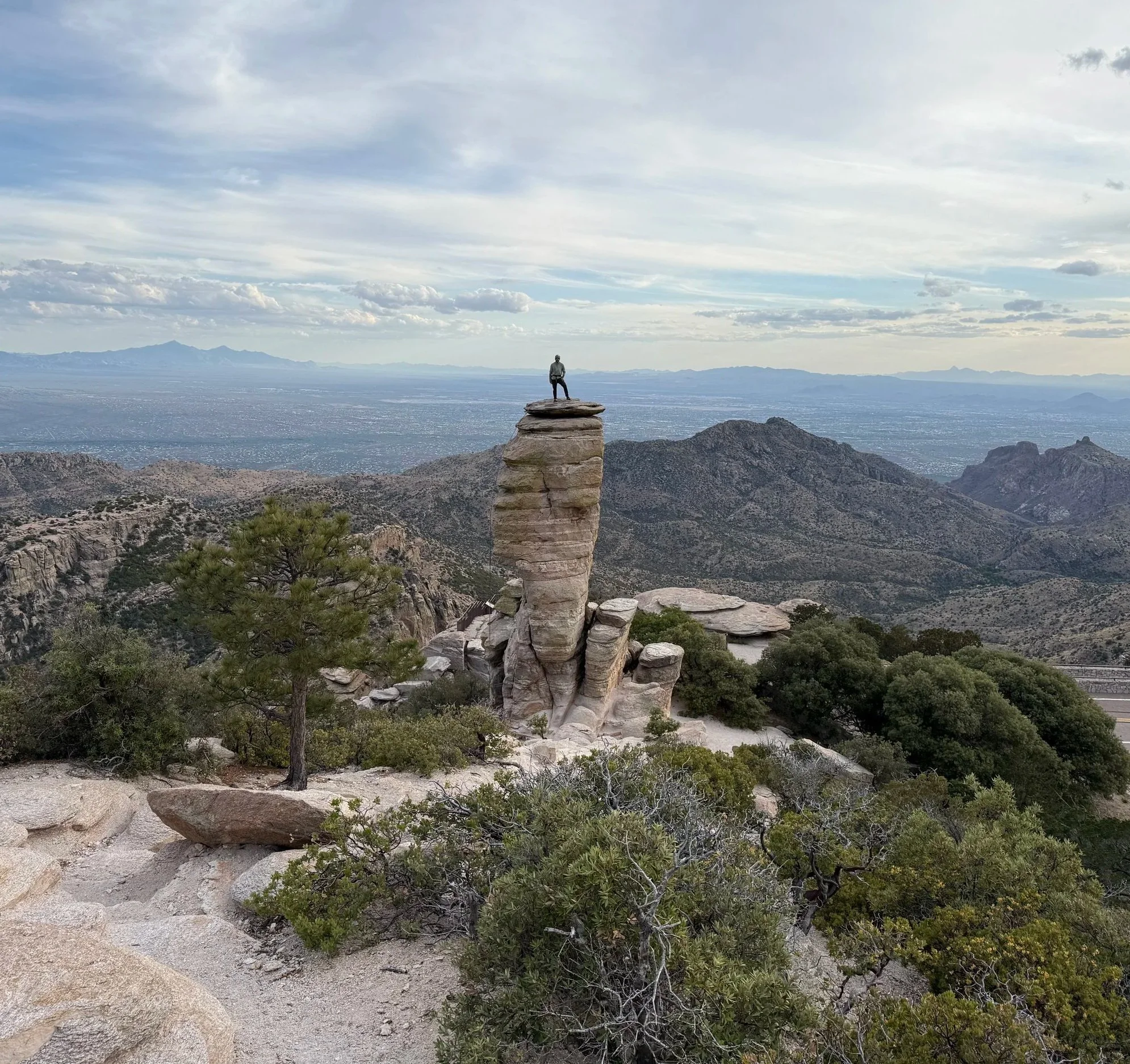 A person standing on top of a large rock formation in a vast desert landscape with mountains in the distance under a partly cloudy sky.