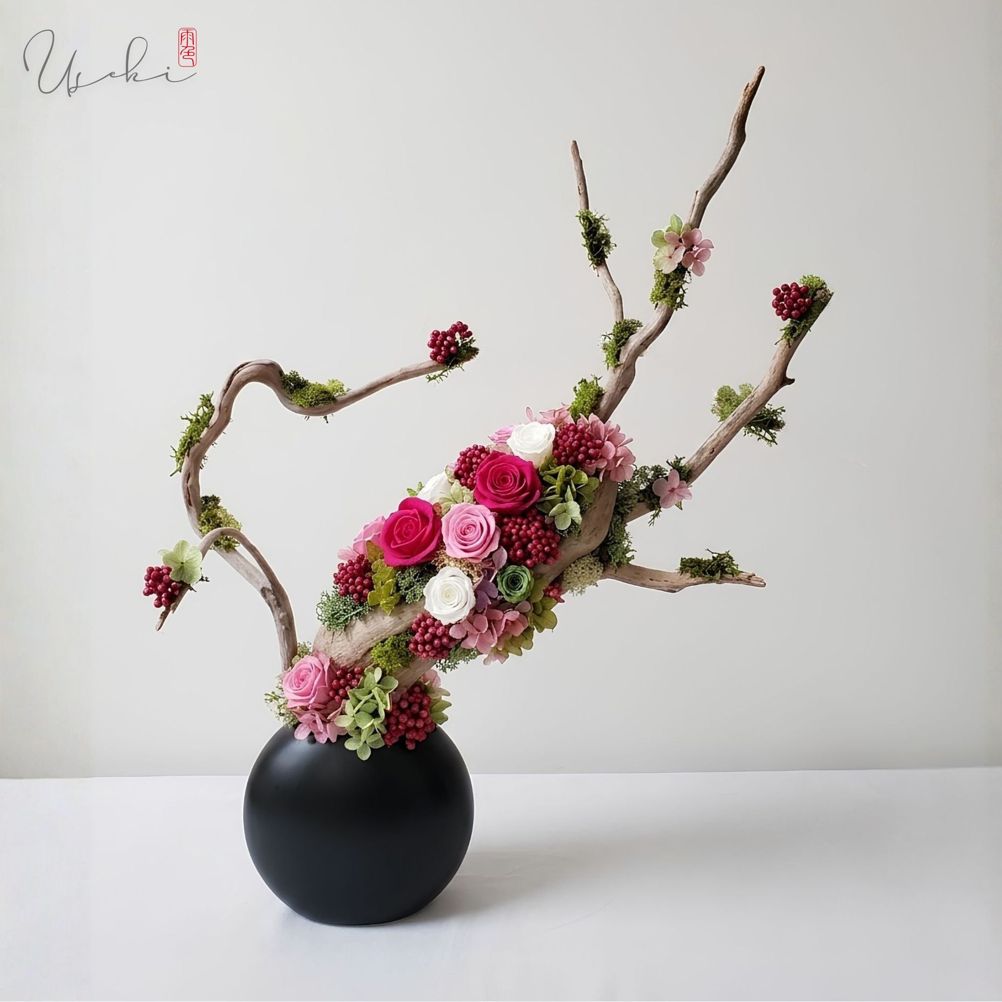 A flower arrangement with pink roses, white roses, purple flowers, red berries, green moss, and branches in a black vase against a white background.