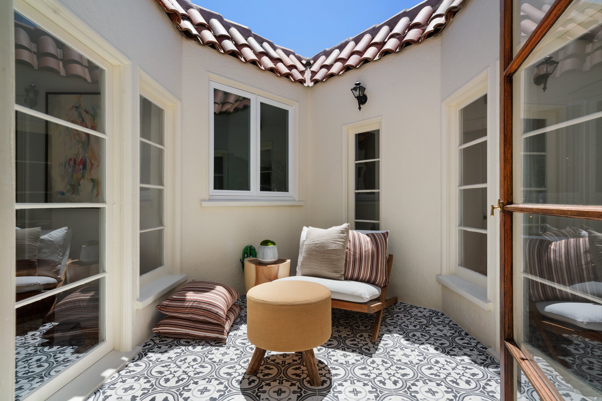 A cozy enclosed patio with white stucco walls, black and white patterned floor tiles, and French doors with glass panes. The patio is furnished with striped and solid cushions, a small round wooden table, and decorative plant pots. A black outdoor wa