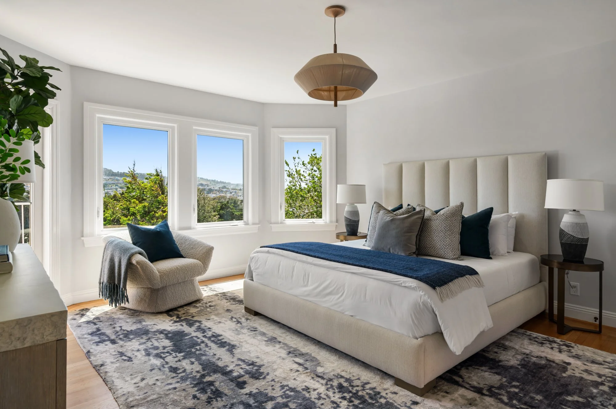 Bright bedroom with large windows showing a scenic view, white upholstered bed with pillows and a dark blue blanket, matching bedside lamps, a cozy chair with a throw blanket, a potted plant, and a patterned area rug.