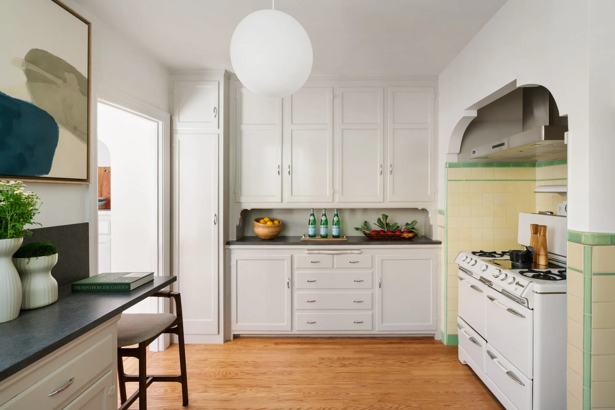 A kitchen with white cabinets, a white stove, and light green tiled backsplash, wooden flooring, and a small counter with decorative plants and a book.