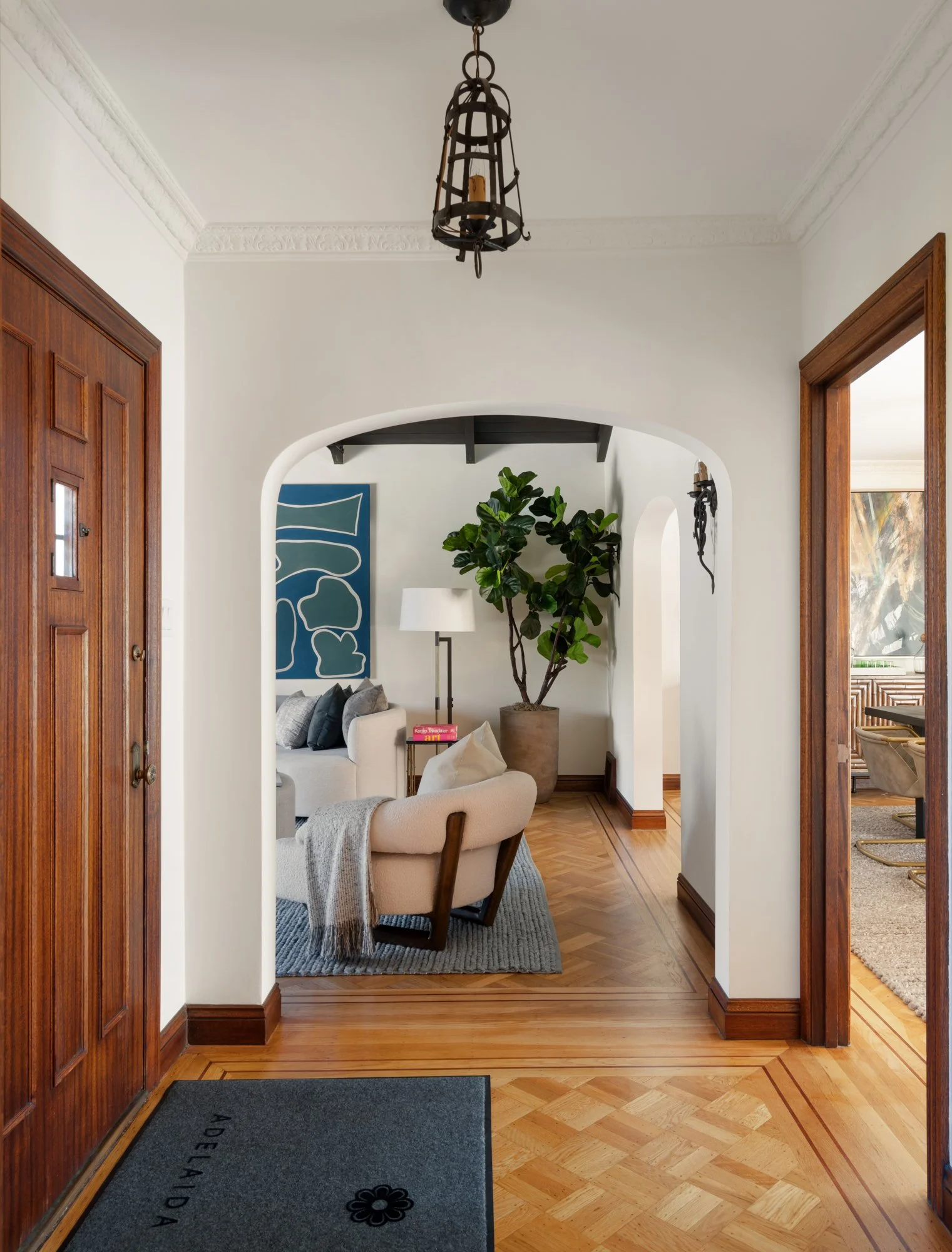 View of the interior of a house featuring an entryway with a wooden door, a decorative wrought iron chandelier, and a living room with white walls, a large potted plant, a navy blue abstract art piece, and a chandelier on the wall. Hardwood flooring 