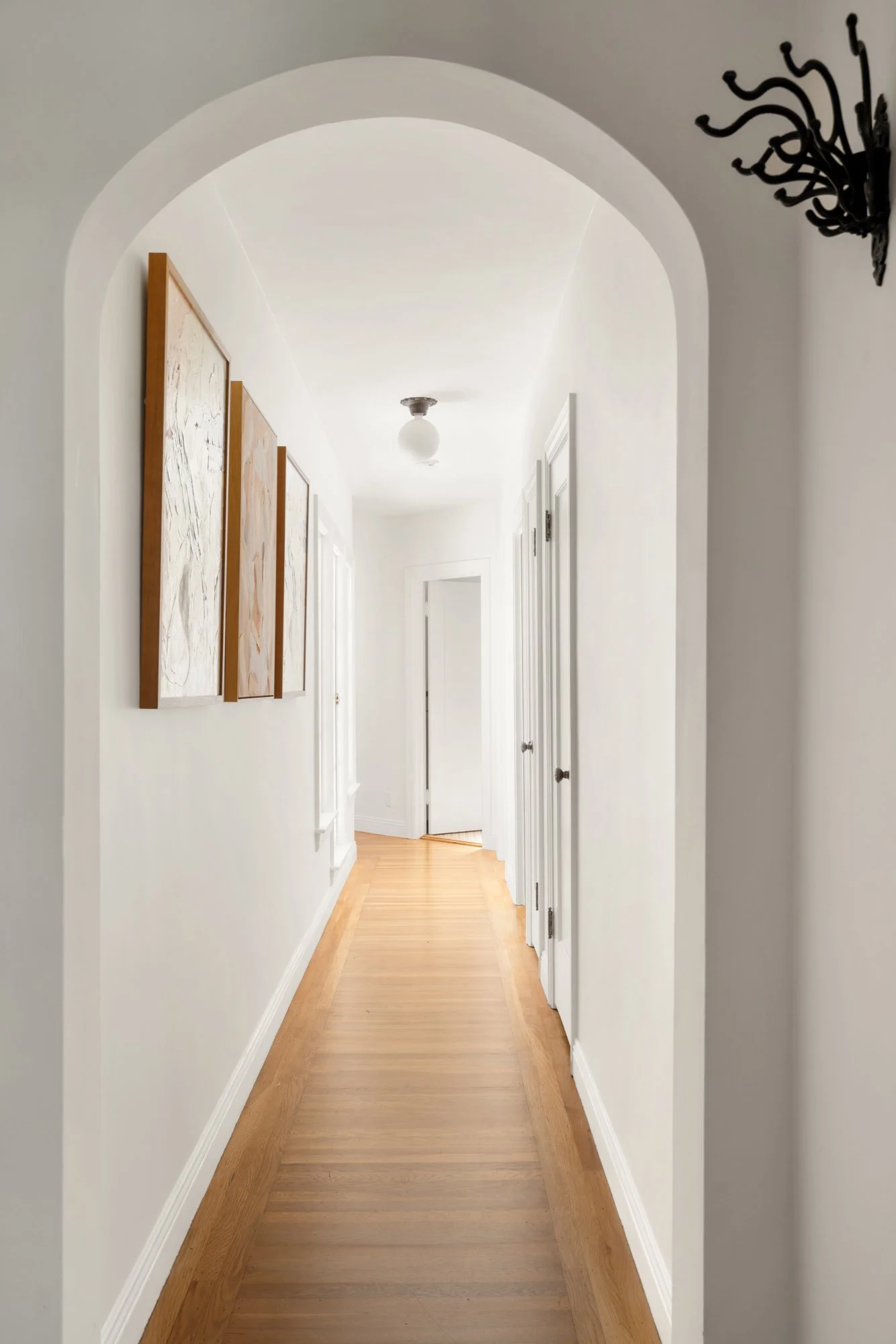 A bright, narrow hallway in a house with white walls, wooden flooring, and multiple white doors on the right side. Artwork hangs on the left wall, and a decorative black wall hook is visible on the top right corner. A ceiling light fixture is at the 