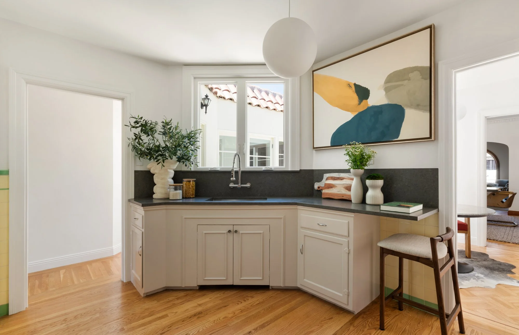 A bright, modern kitchen with white cabinets, a black countertop, and a window. Decor includes potted plants, jars, a chopping board, and a large abstract wall art piece. Part of a dining area is visible to the right.